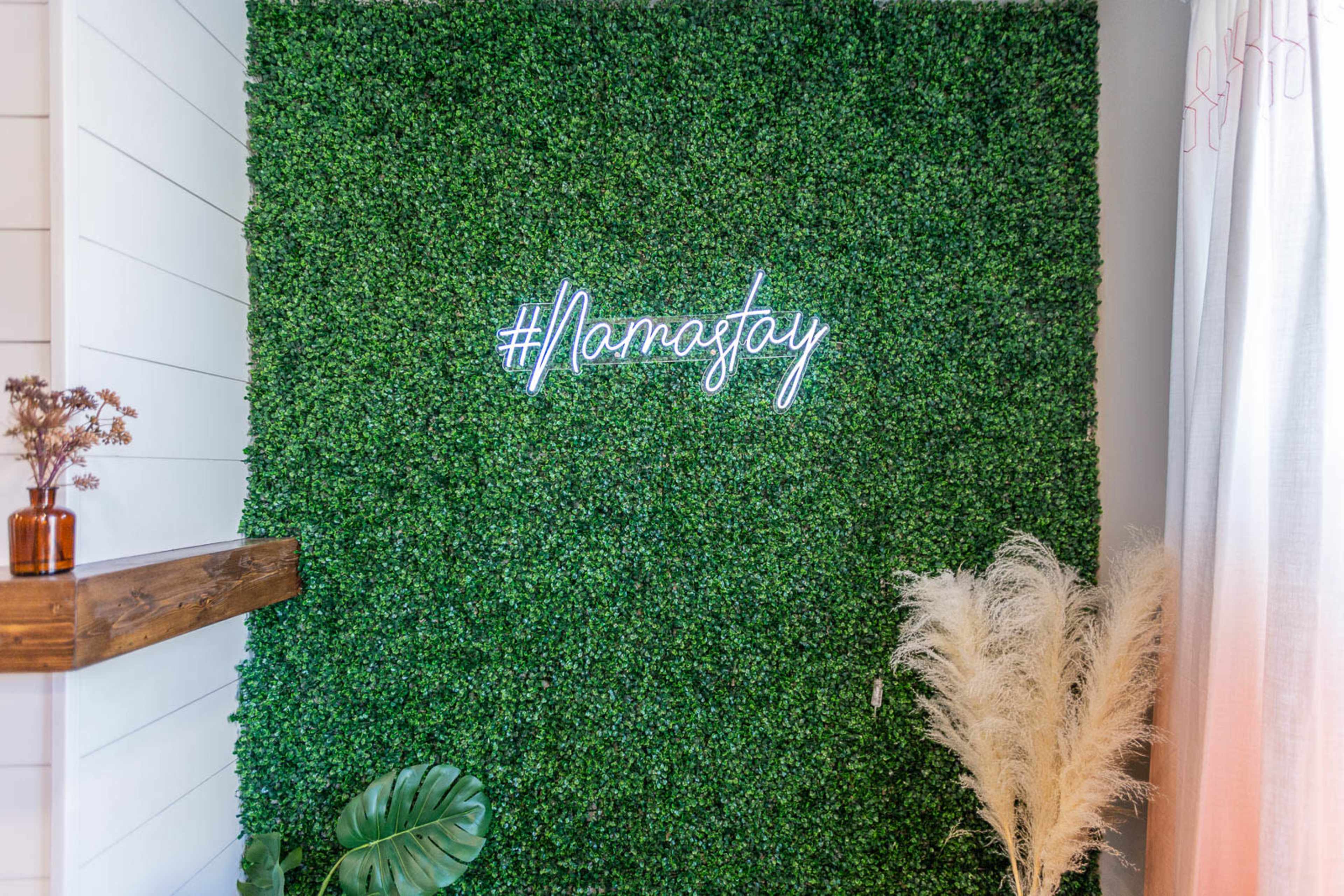 A green wall made of artificial foliage with a neon sign that reads "#Namaslay" at the center.