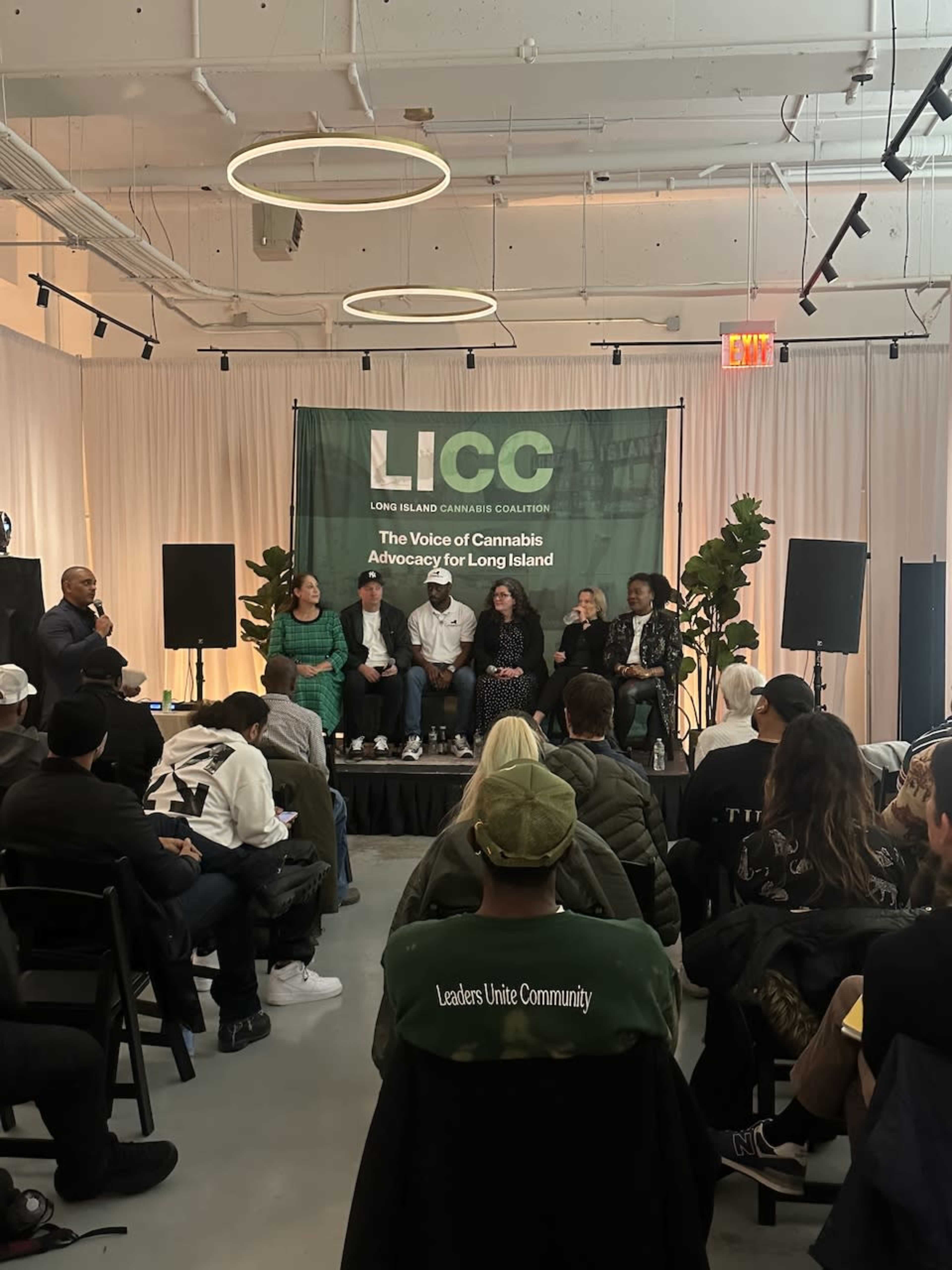 A panel discussion takes place in a well-lit room with a group of speakers seated on a stage, while attendees listen and engage from seating arranged in front of them.