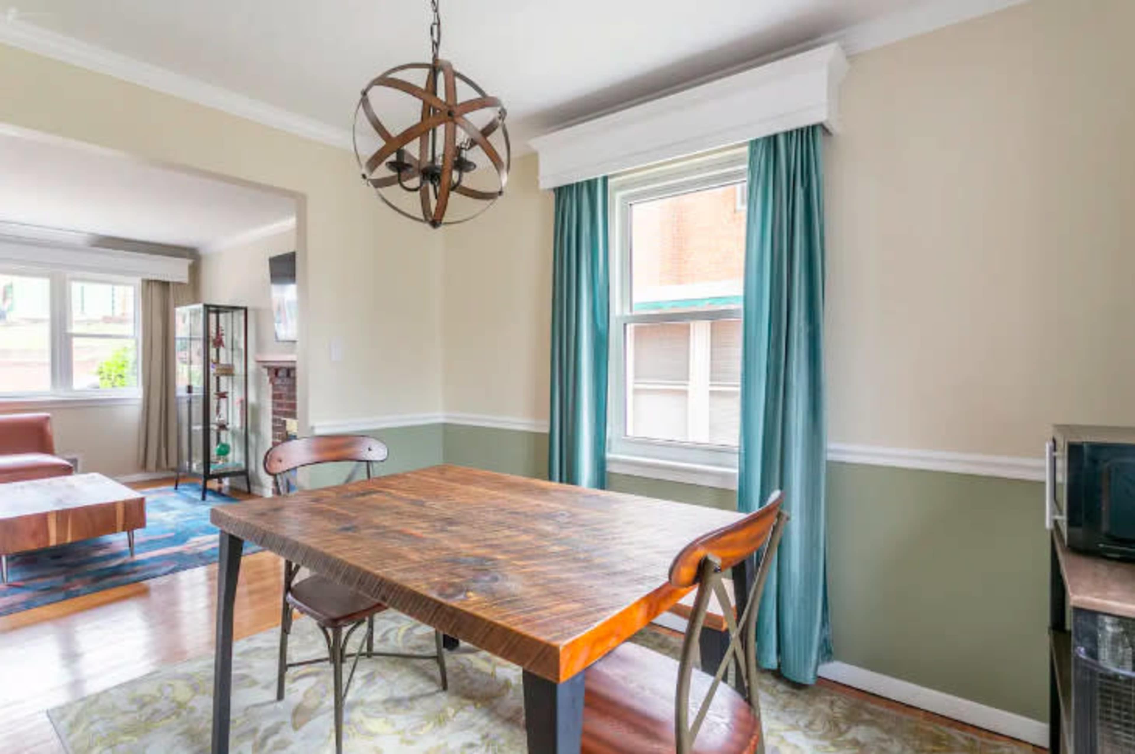 The image shows a dining area with a wooden table and chairs, a window dressed with blue curtains, and a decorative light fixture overhead.