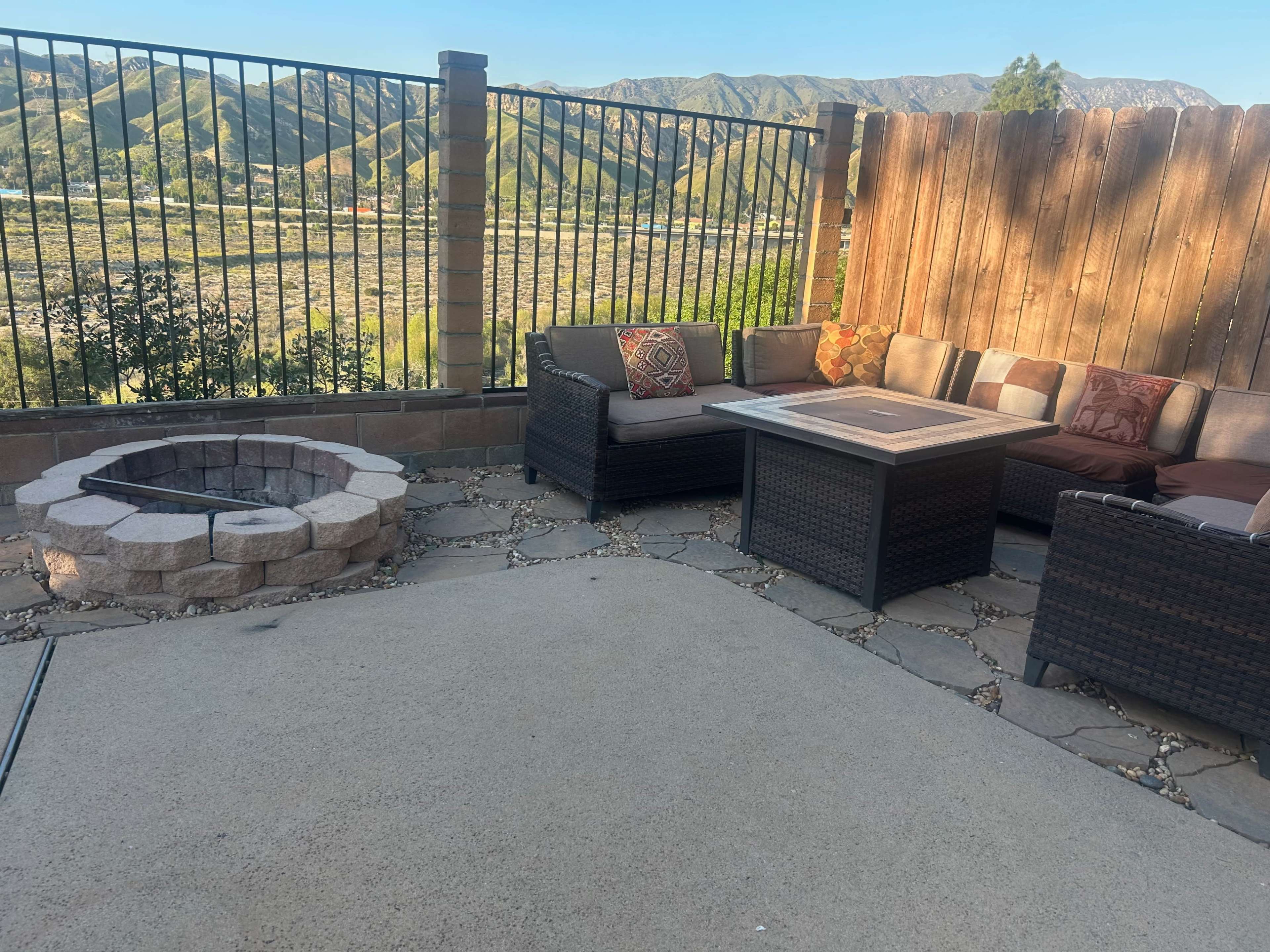 A patio area features a stone fire pit, a table, and wicker seating surrounded by a fence with mountains in the background.