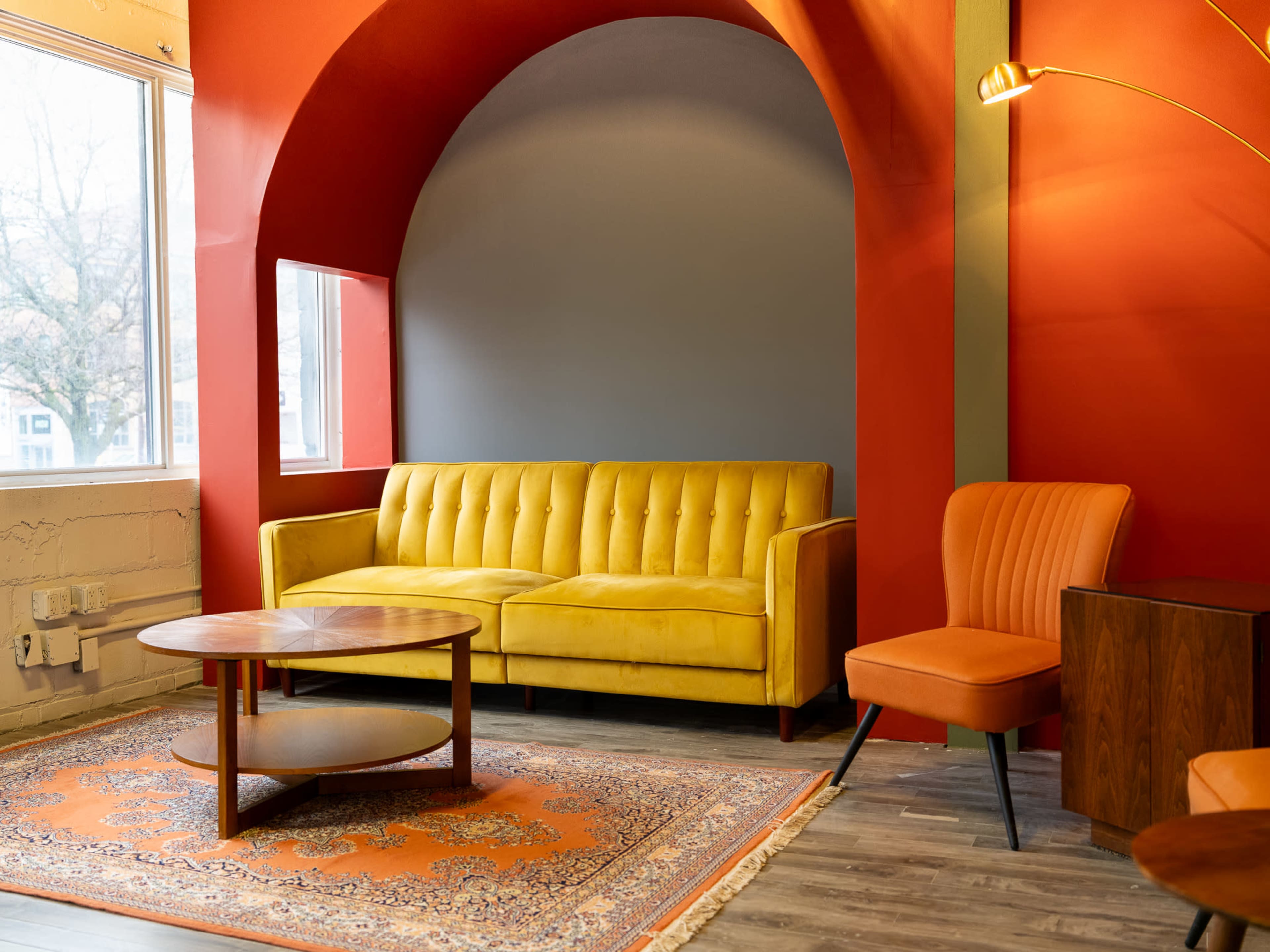The scene features a bright, modern interior with a yellow couch, an orange armchair, a round wooden coffee table, and a patterned area rug against a red and gray wall.