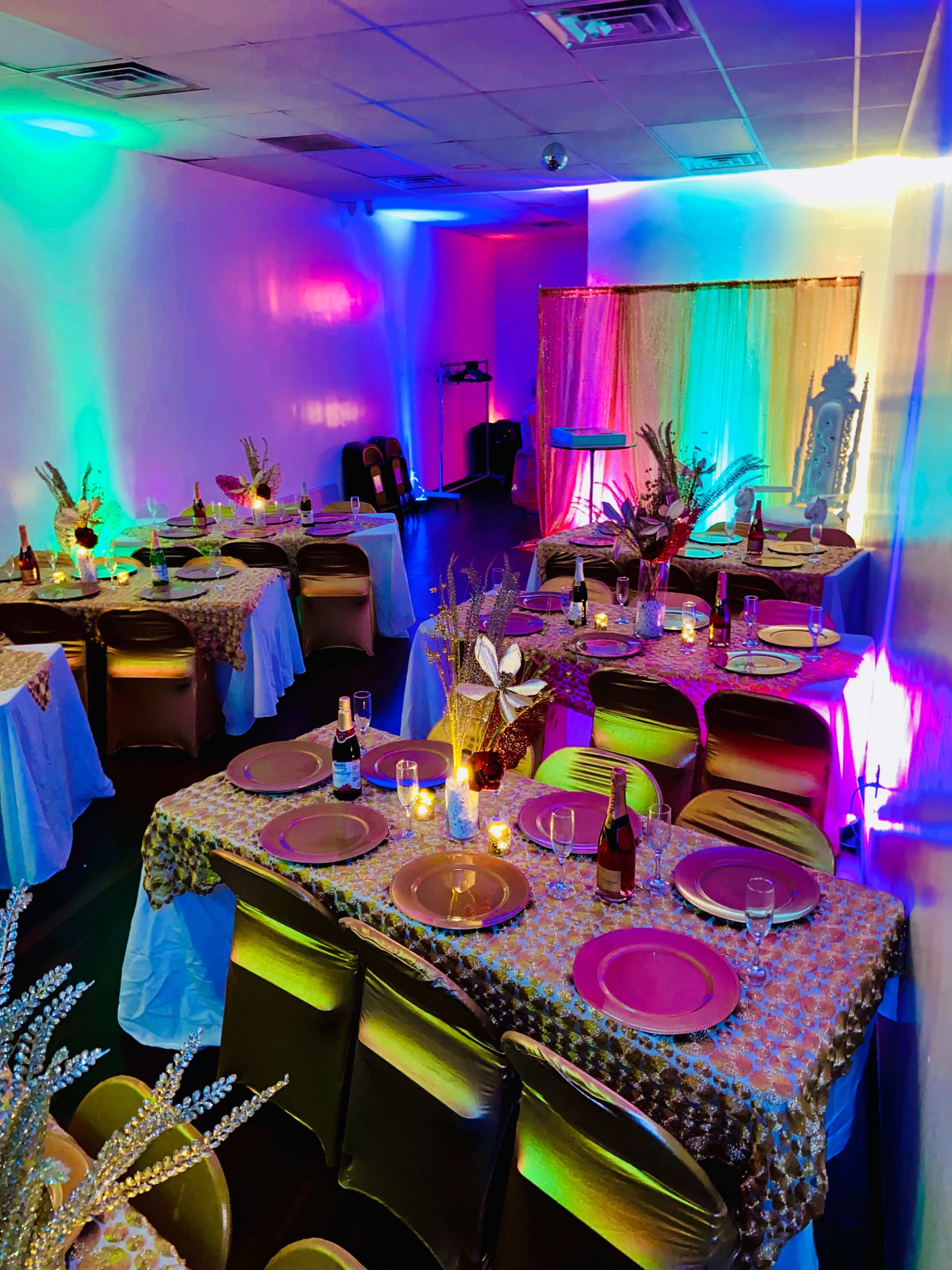 A banquet room with tables set for a celebration, featuring colorful lighting and decorative centerpieces.