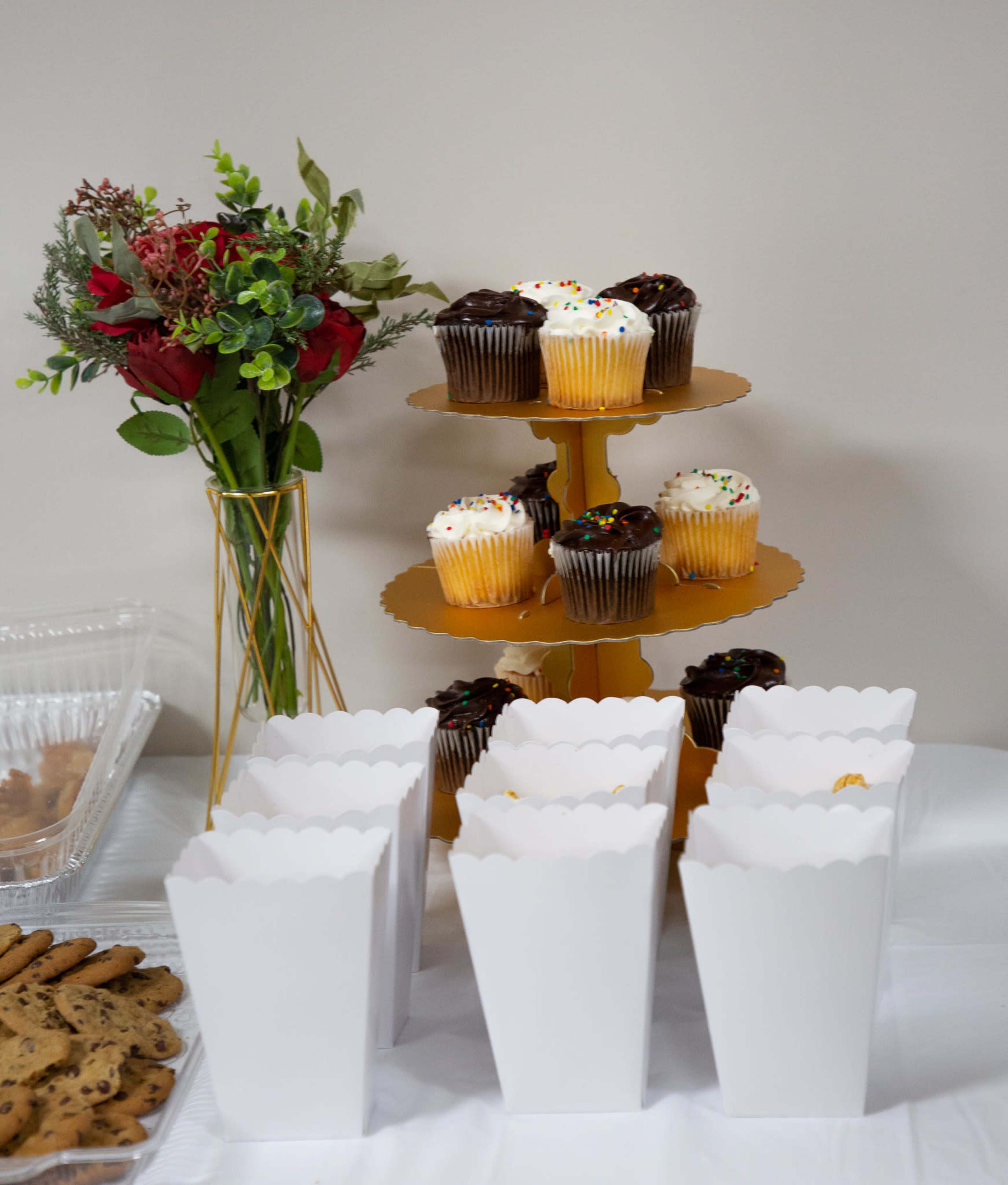A tiered cake stand holds colorful cupcakes, while white paper boxes are lined up in front, and a bouquet of flowers adds a decorative touch to the scene.