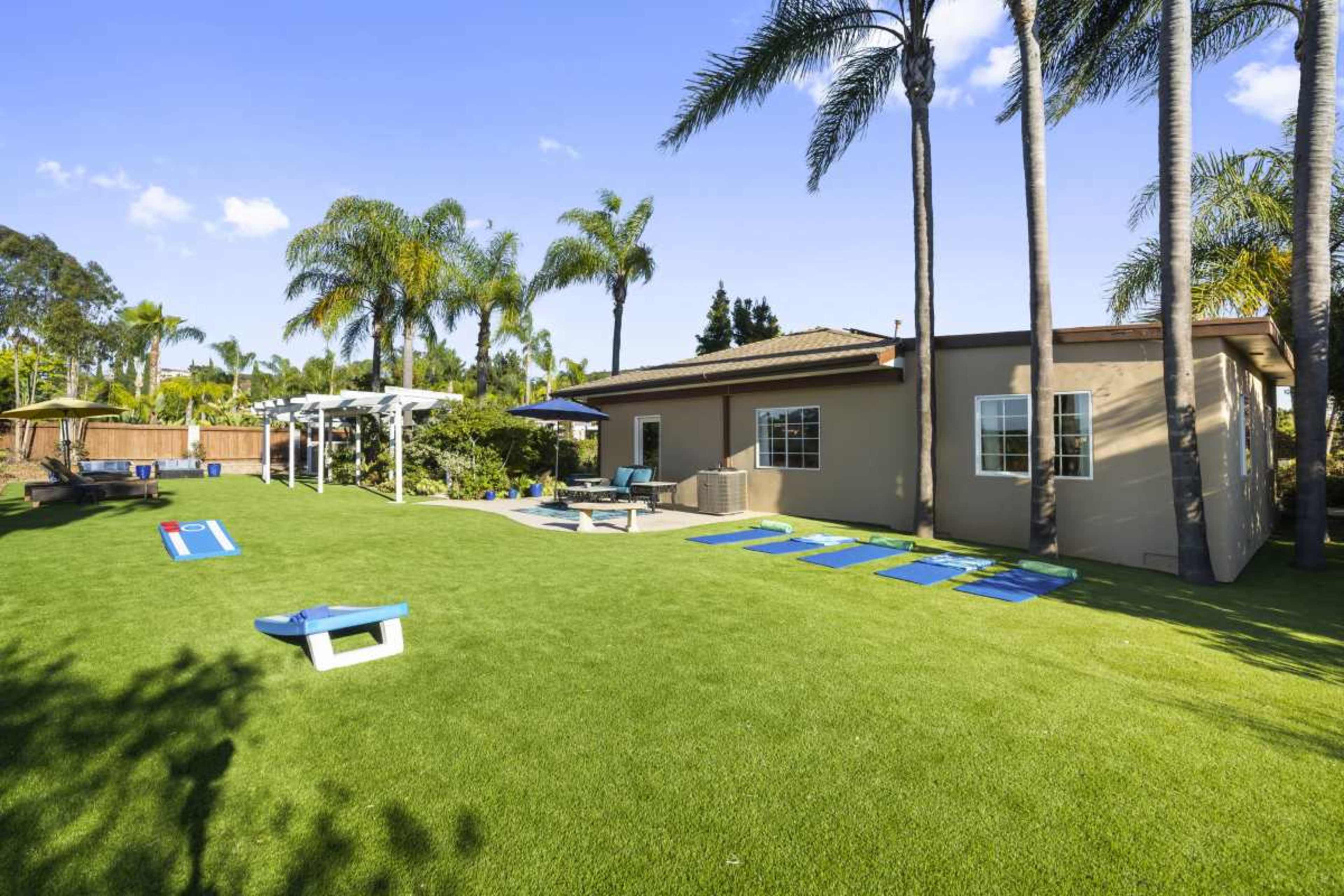 The image shows a spacious backyard with green grass, palm trees, lounge chairs, and a swimming pool beside a house.