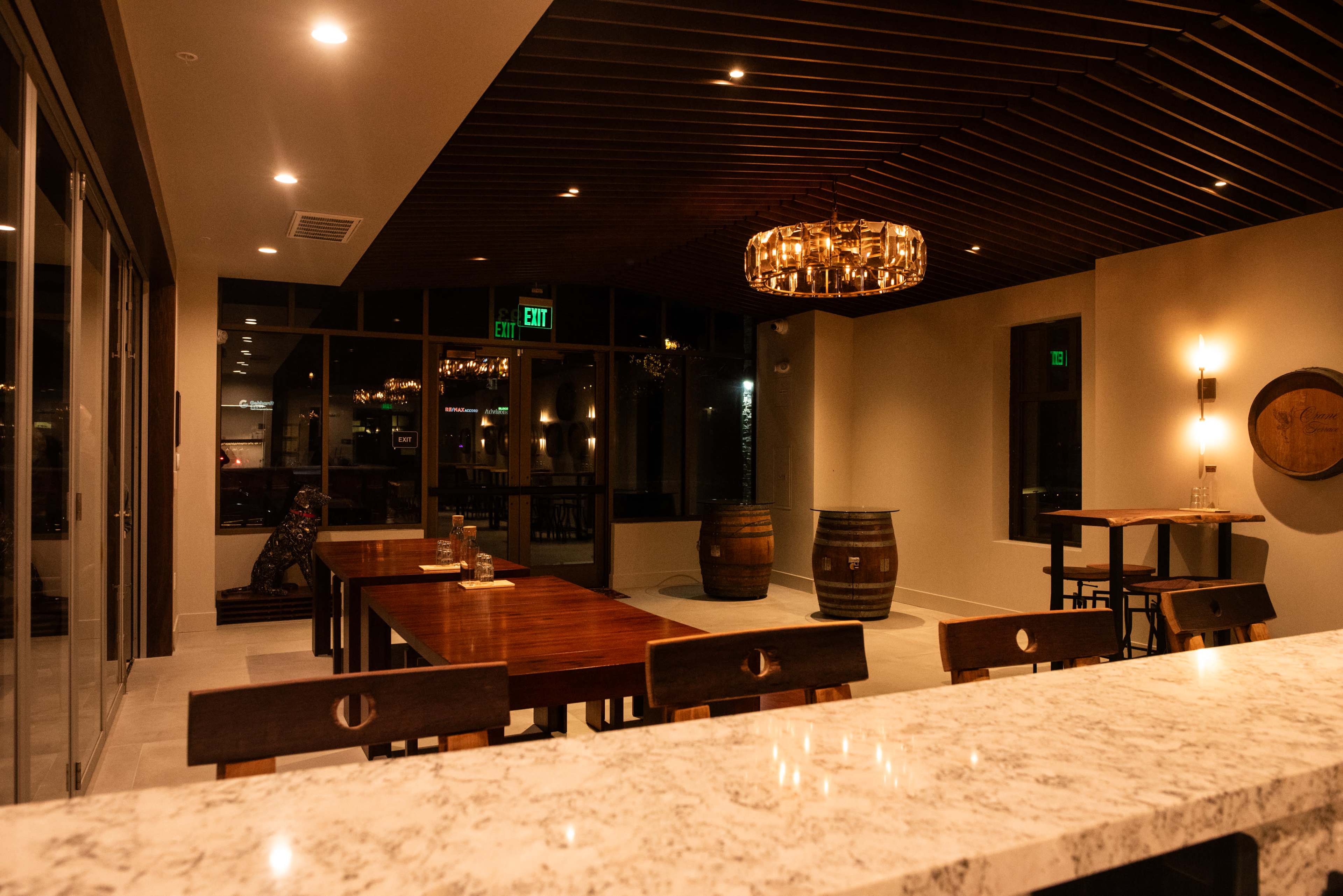 The image shows a modern, upscale interior of a restaurant or bar with wooden tables, barrel-style decor, and a marble countertop illuminated by warm lighting.