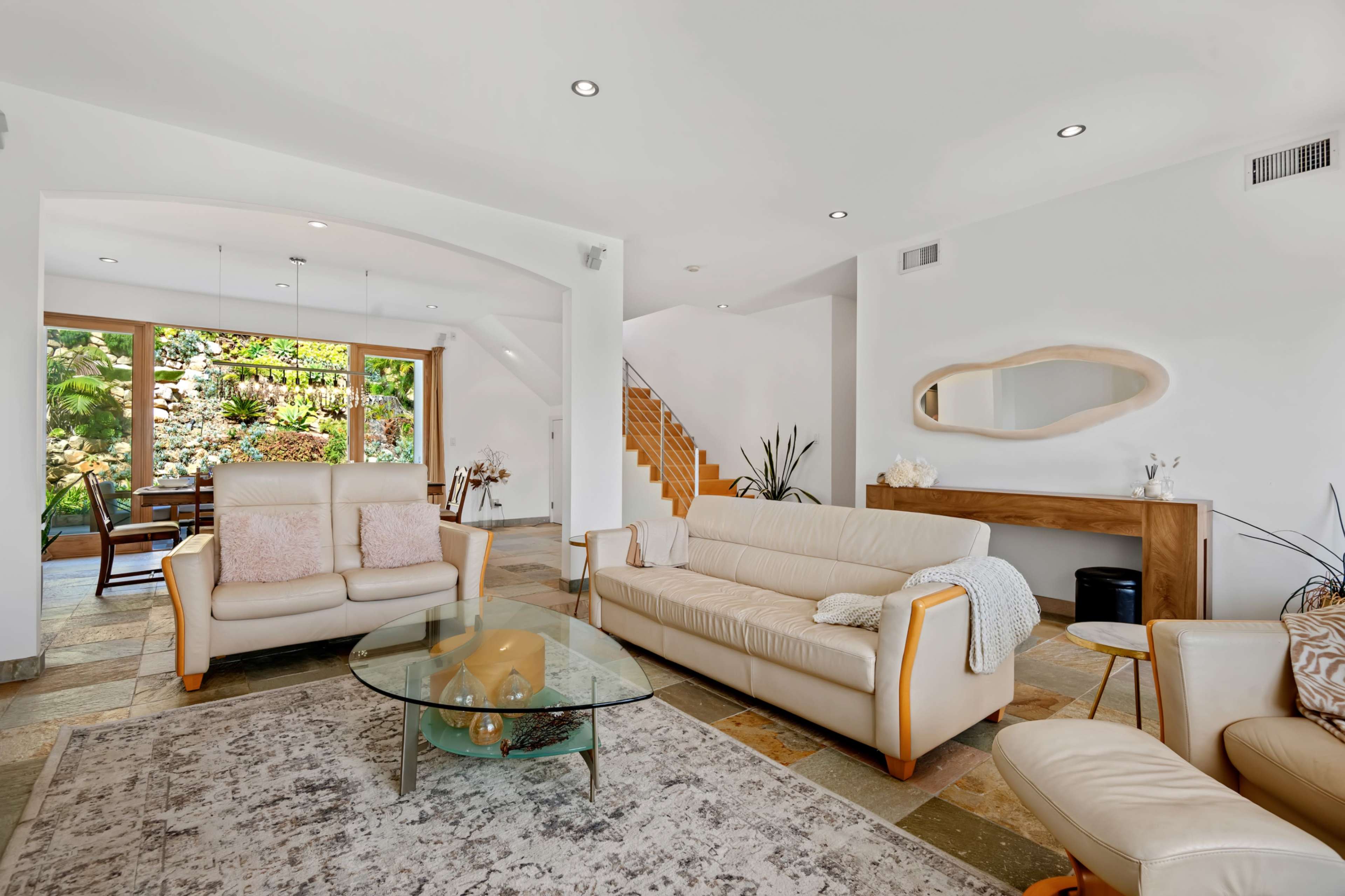 The living room features a beige leather sofa set, a glass coffee table, and a large window overlooking a garden.