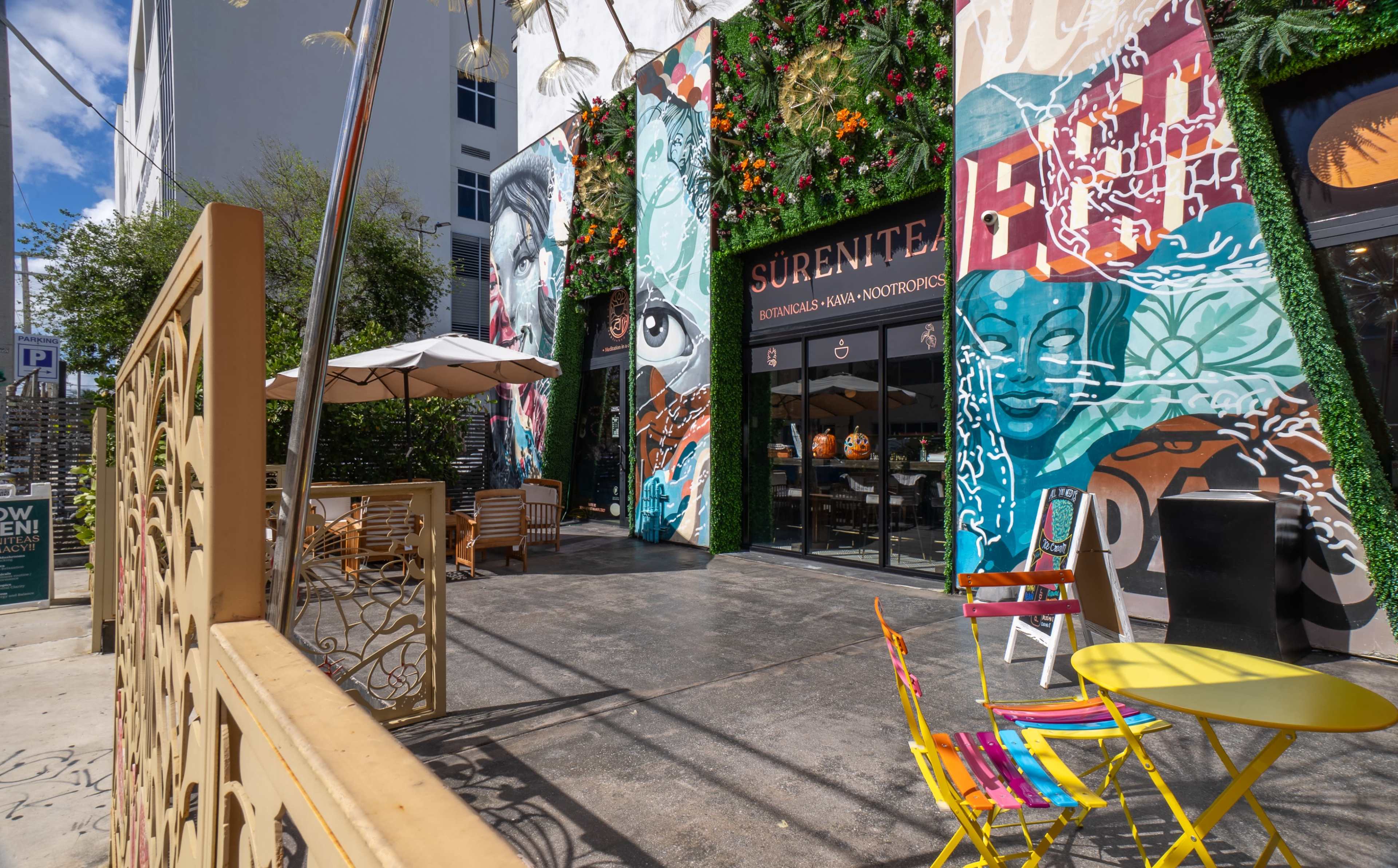 The scene features a vibrant café front with colorful murals on the exterior walls and outdoor seating in a sunny urban setting.