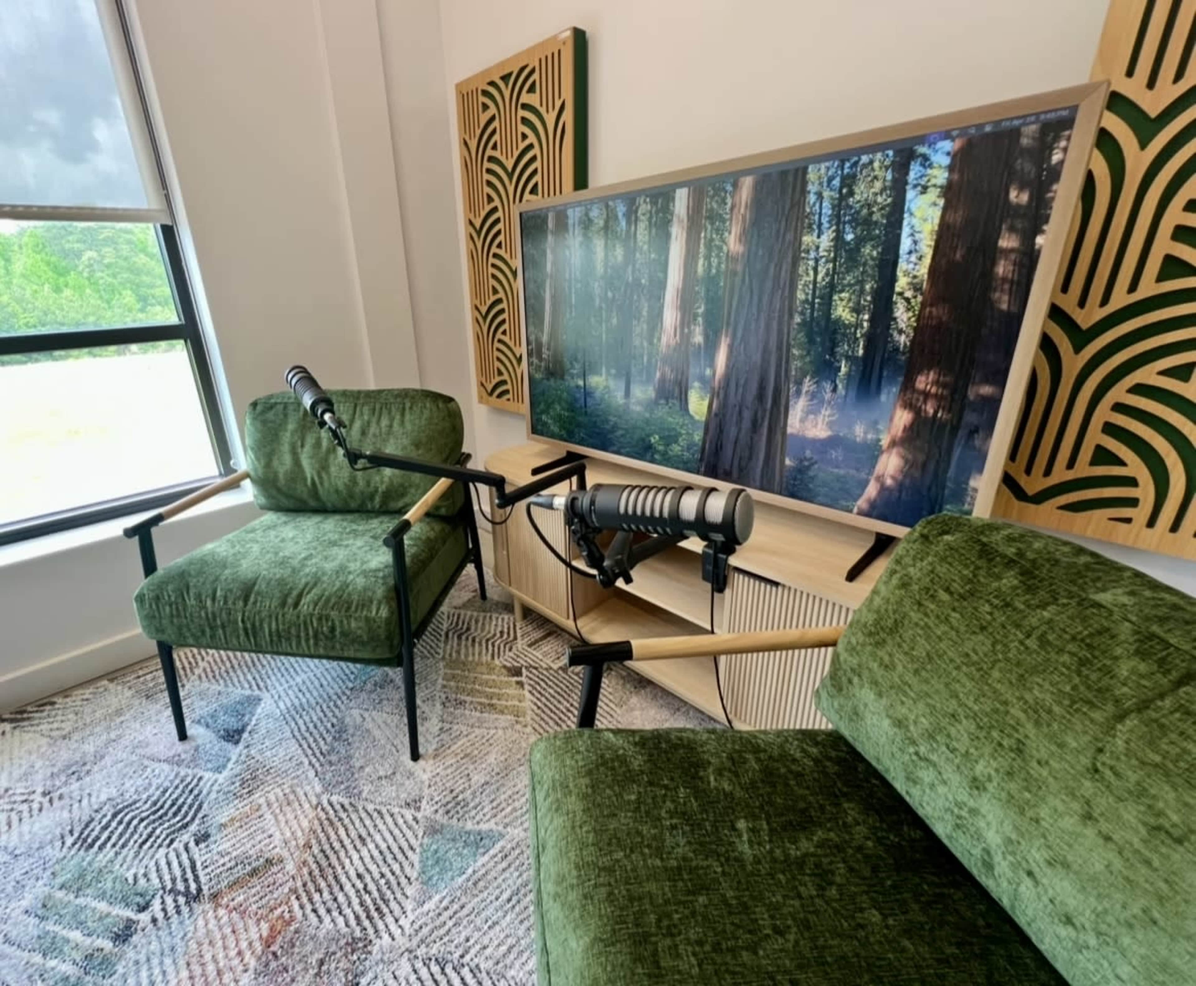 A cozy recording space features two green chairs, a large screen displaying a forest scene, and a microphone setup on a wooden table.