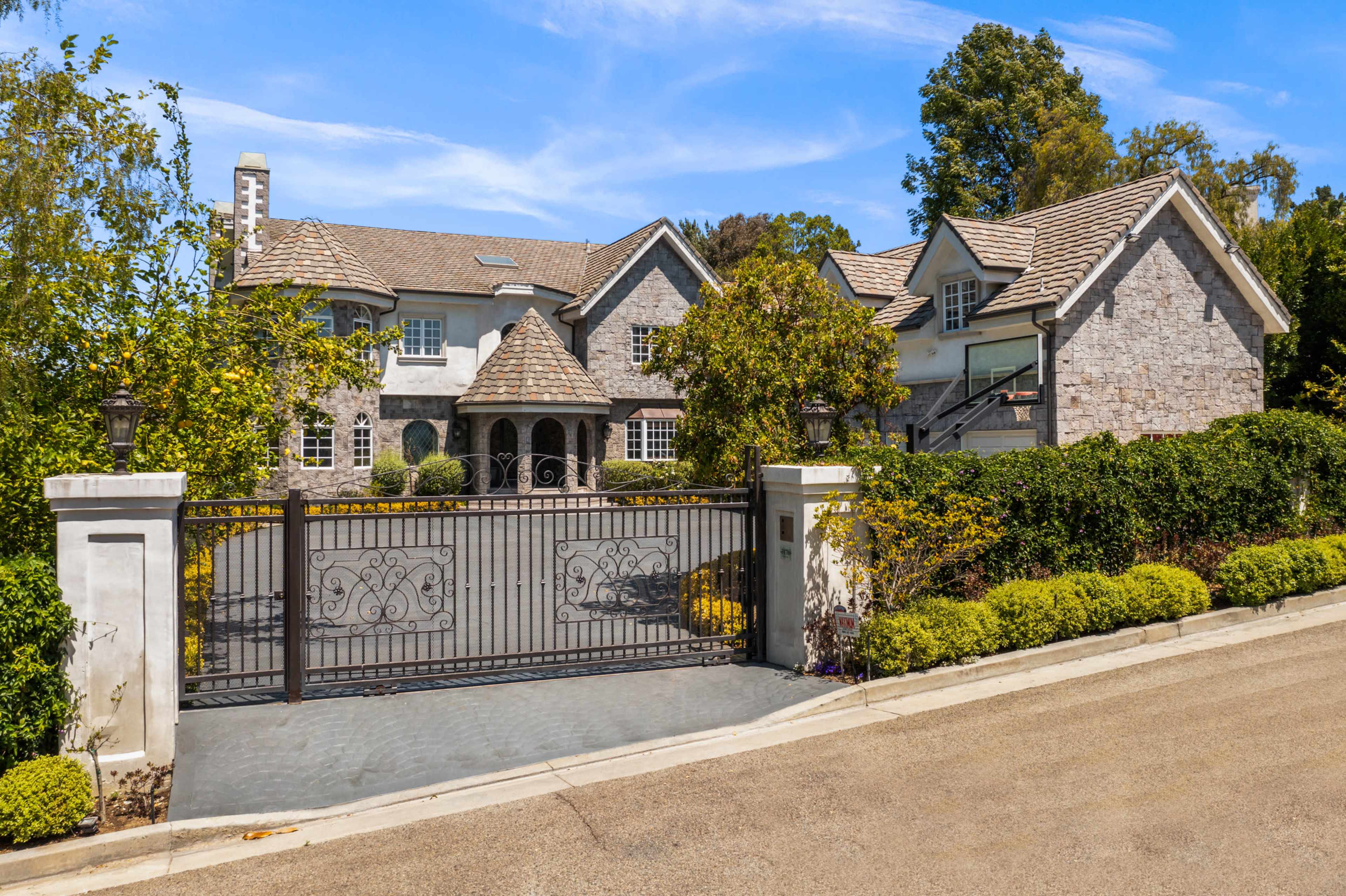 A large stone house with a tiled roof is set behind an ornate metal gate, surrounded by lush greenery and neatly trimmed hedges.