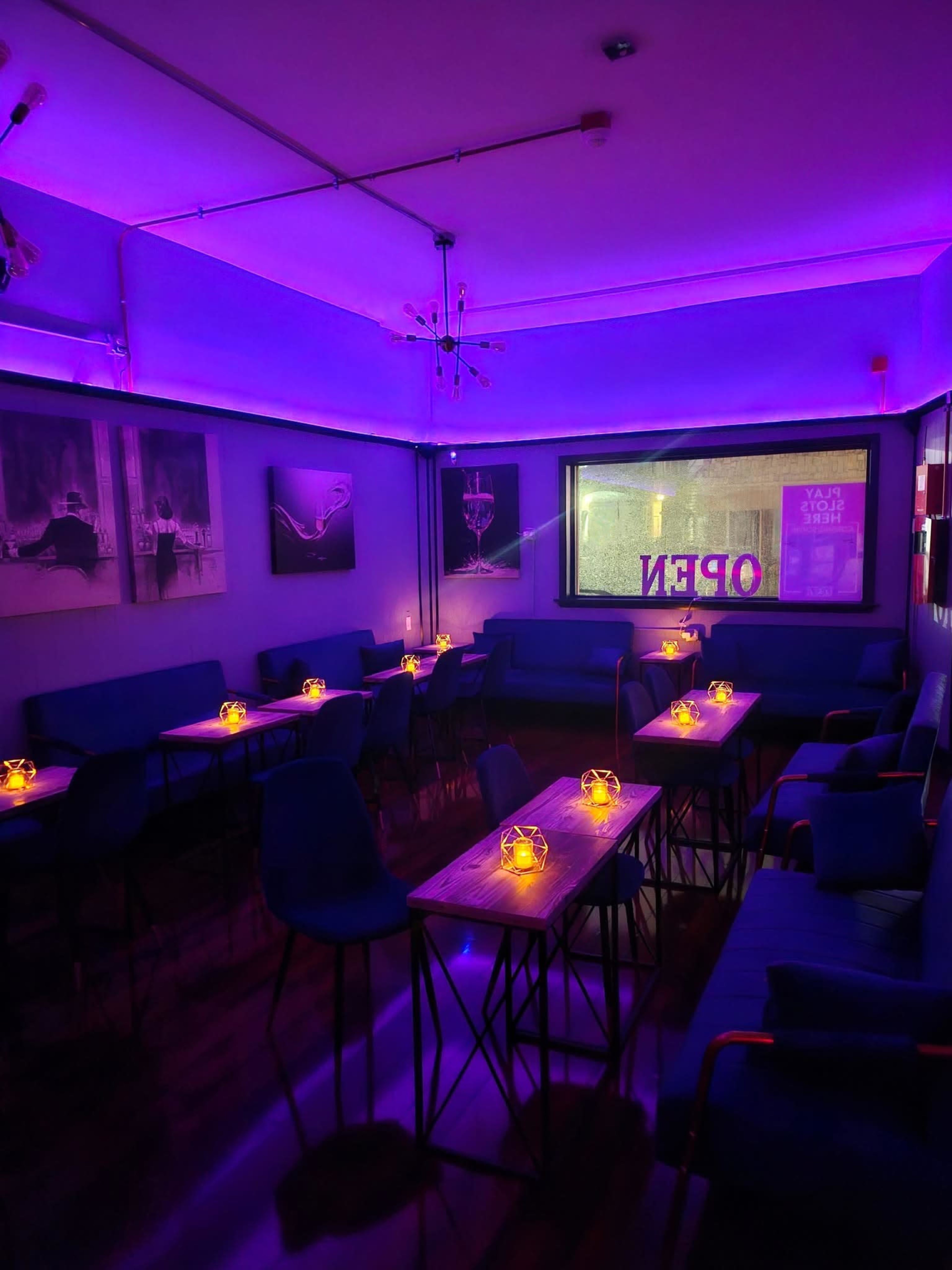 The image shows a dimly lit lounge with purple lighting, featuring tables adorned with candle holders and chairs set up for guests.