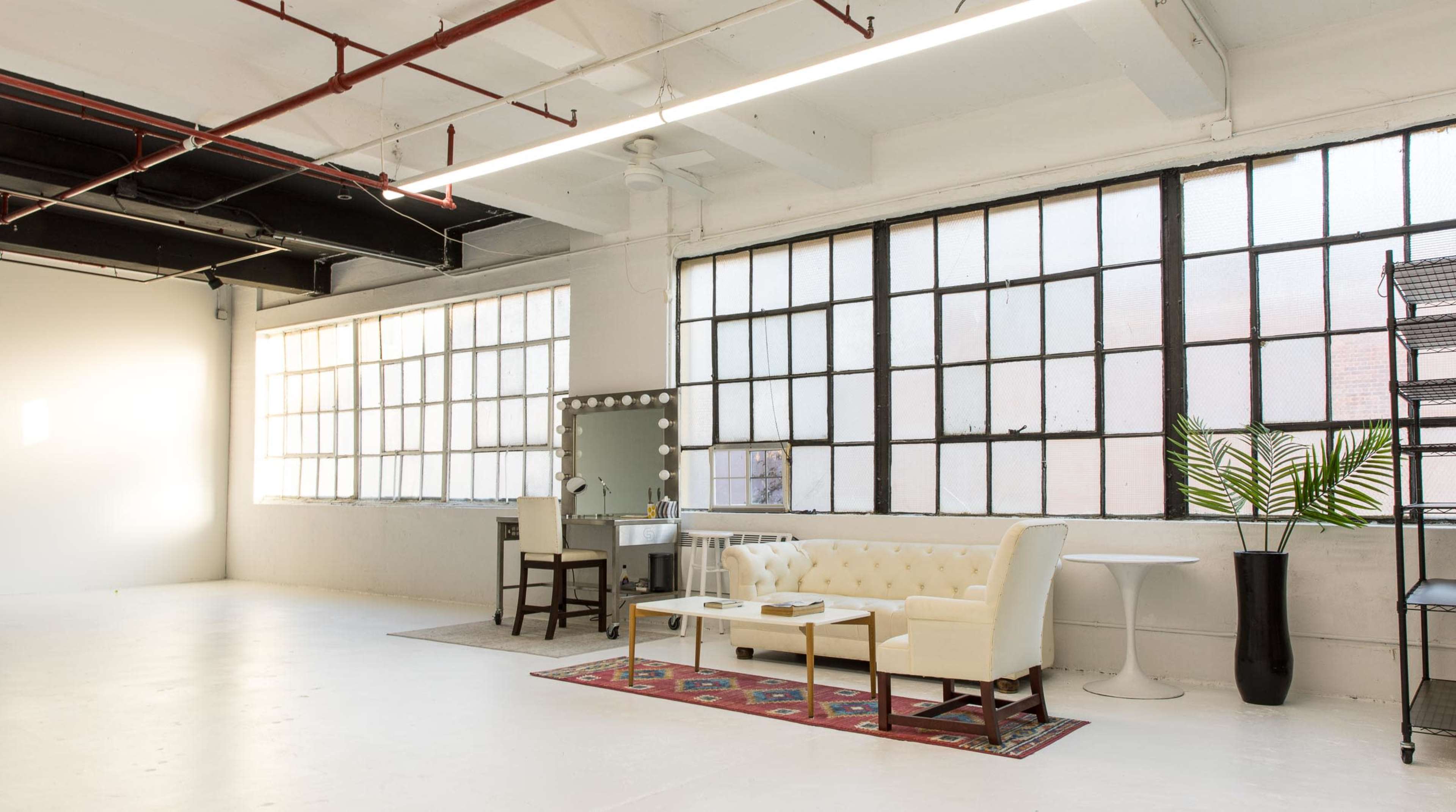 A bright, industrial-style room featuring large windows, a white sofa, a round table, and a decorative plant.