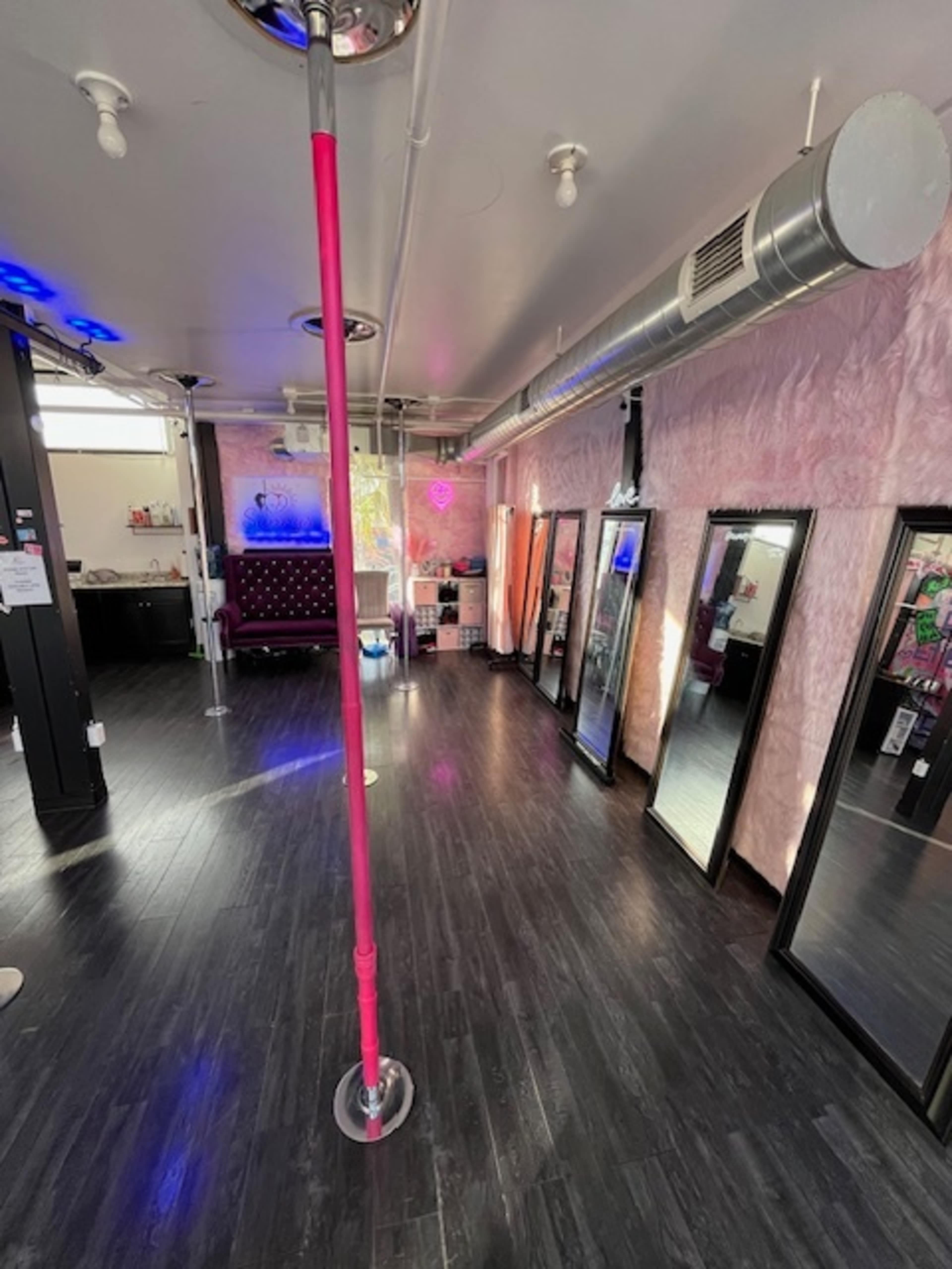 The image shows an interior of a studio with a pink pole in the center, surrounded by several mirrors and decorative lighting.