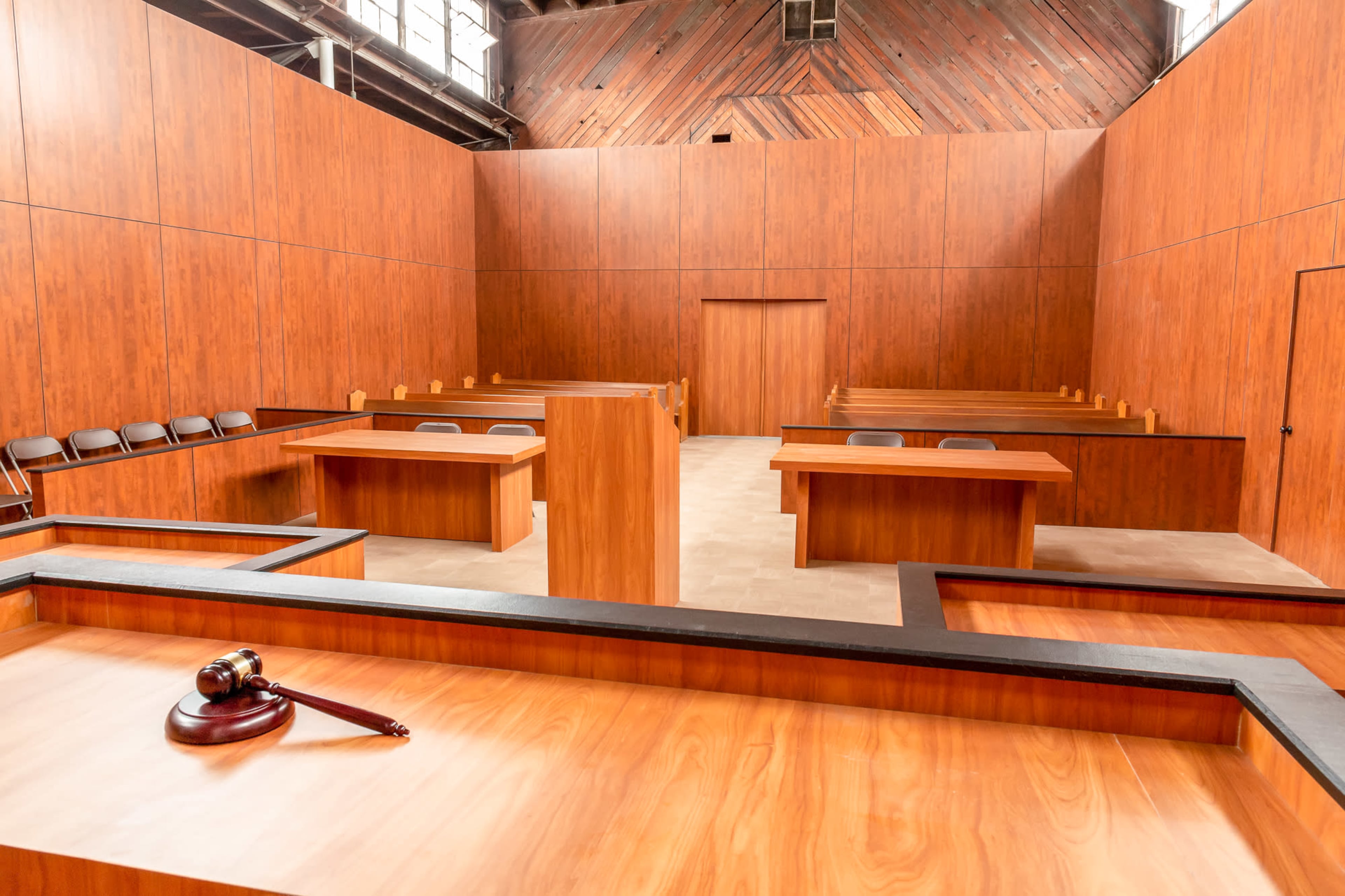 An empty courtroom with wooden paneling, several tables arranged for participants, and rows of chairs.