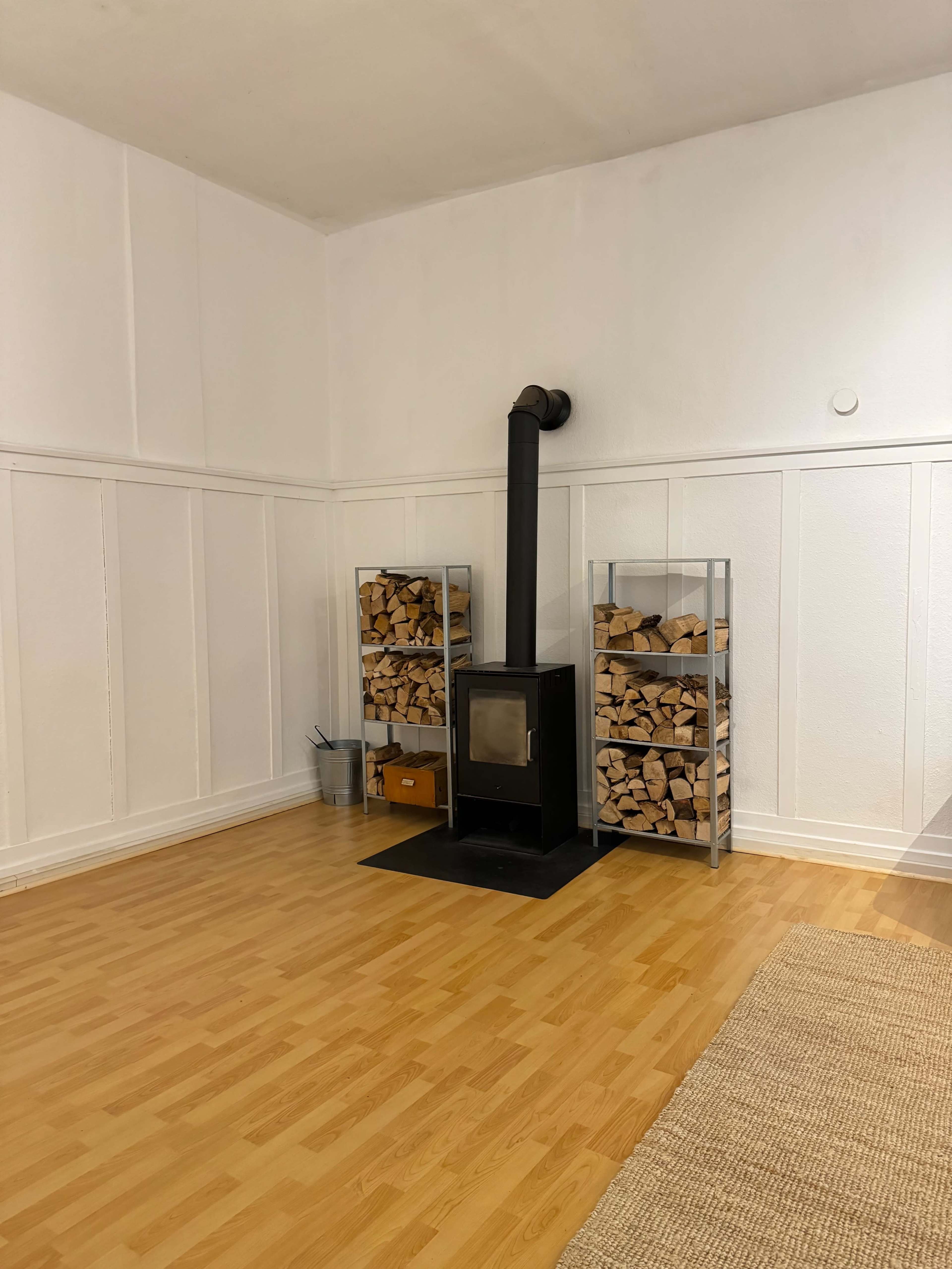 The image shows a minimalistic room featuring a wood-burning stove with two metal racks stacked with firewood on either side.