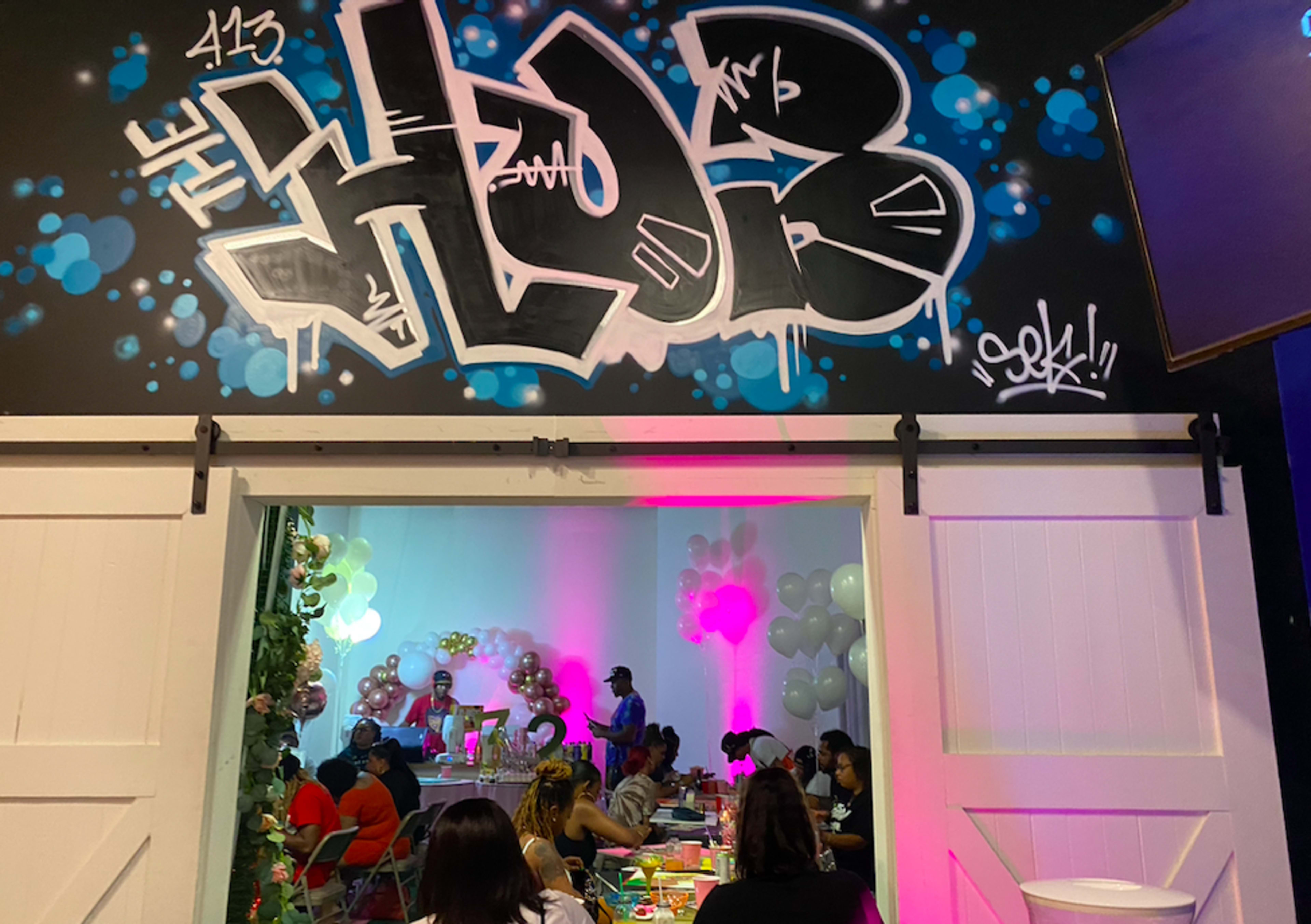A colorful graffiti mural reading "THE HUB" adorns the wall above a gathering of people seated at tables decorated with balloons and party supplies.