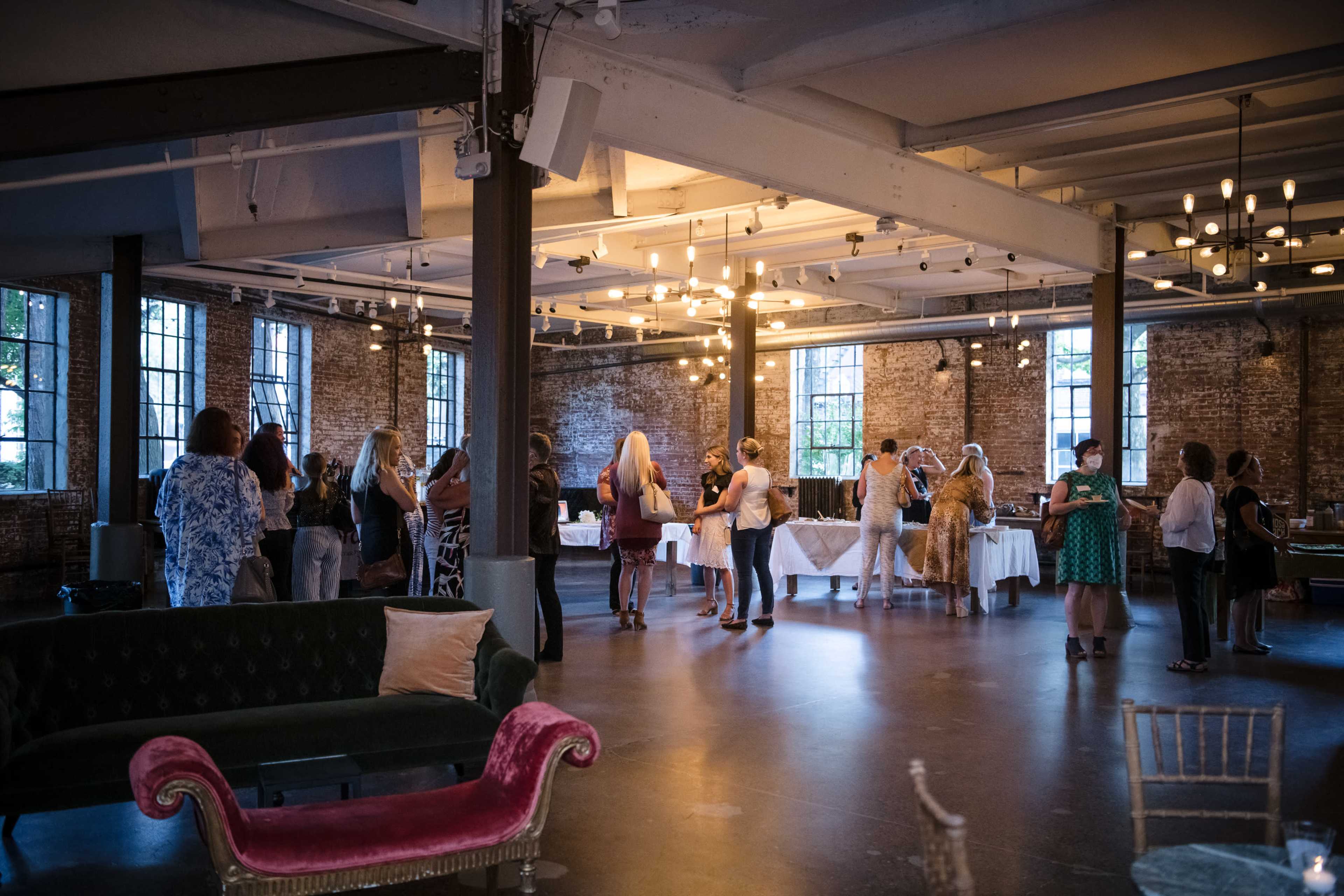 A group of people gathers in a spacious, well-lit event venue with large windows and exposed brick walls.