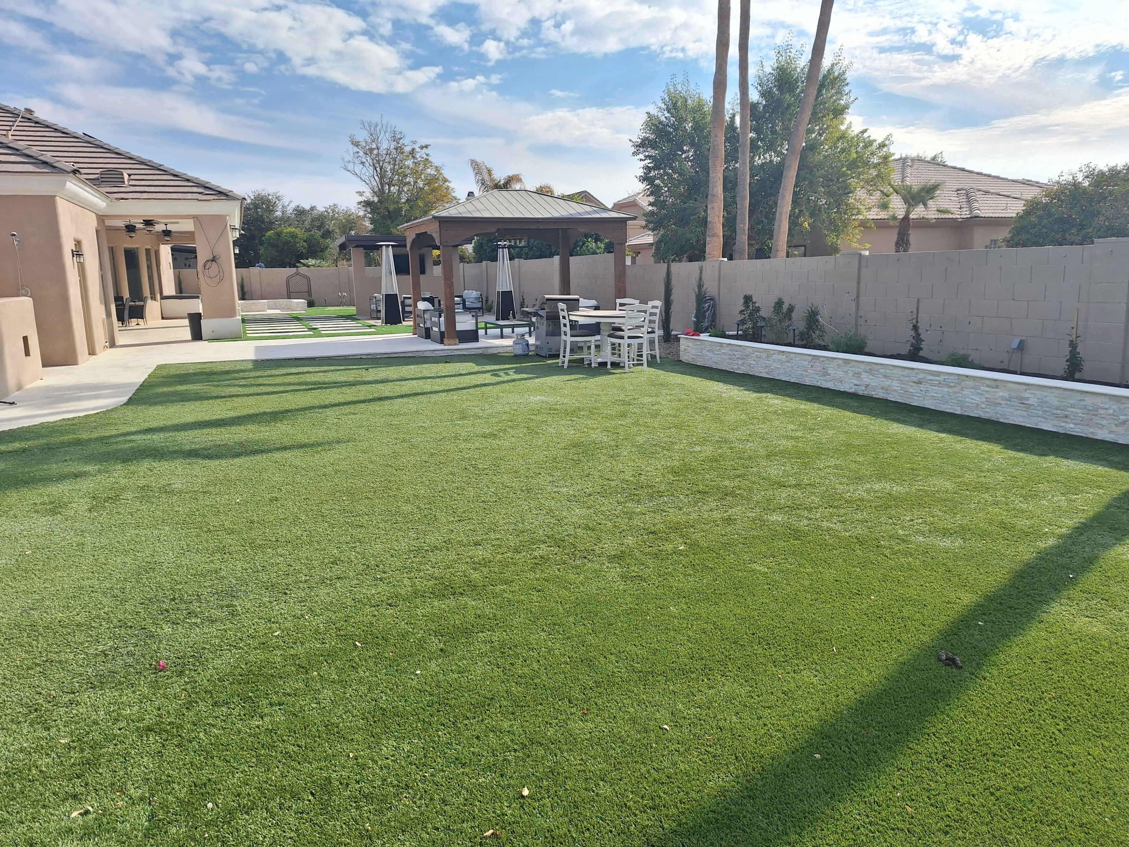 The image shows a landscaped backyard with artificial grass, a gazebo, and seating areas surrounded by trees and a low wall.