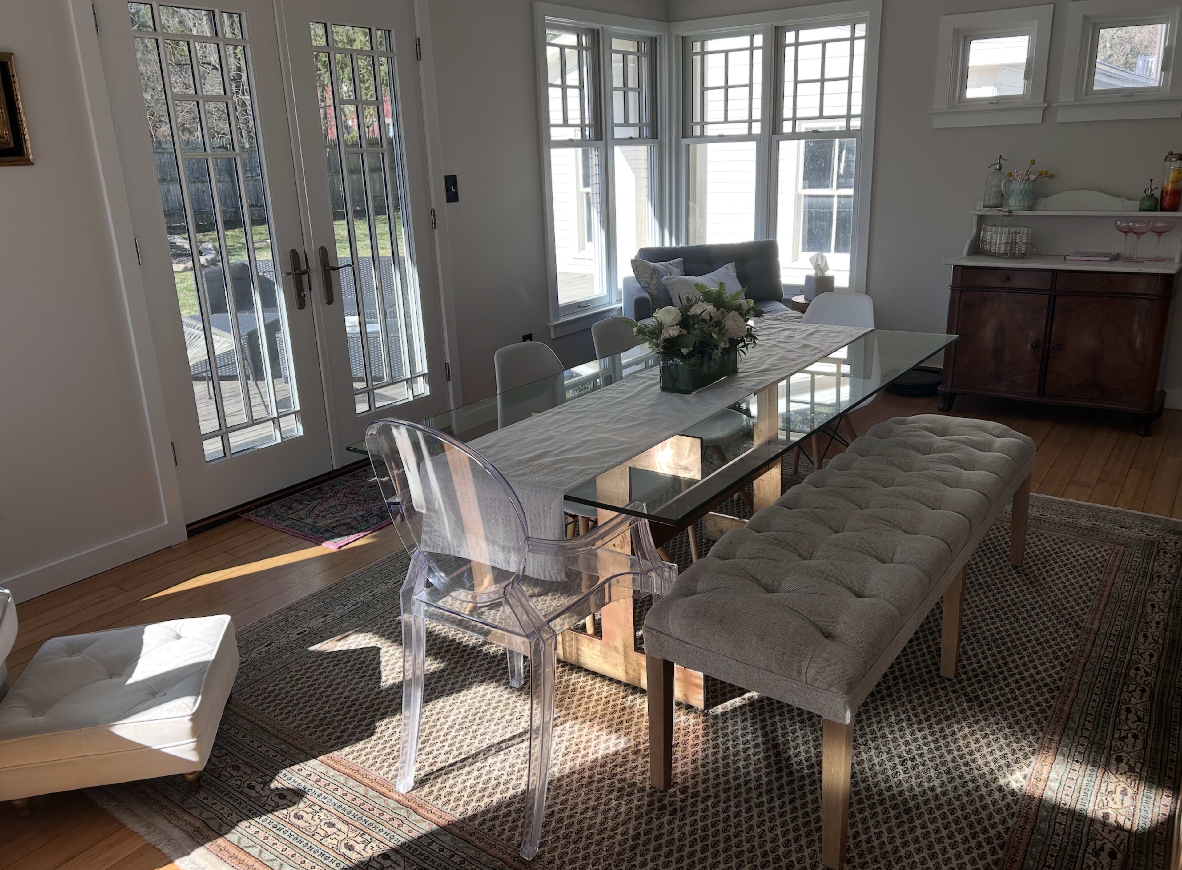 The image shows a well-lit dining area featuring a glass dining table surrounded by transparent chairs, a tufted bench, and a sideboard, with large windows providing a view of the outdoors.