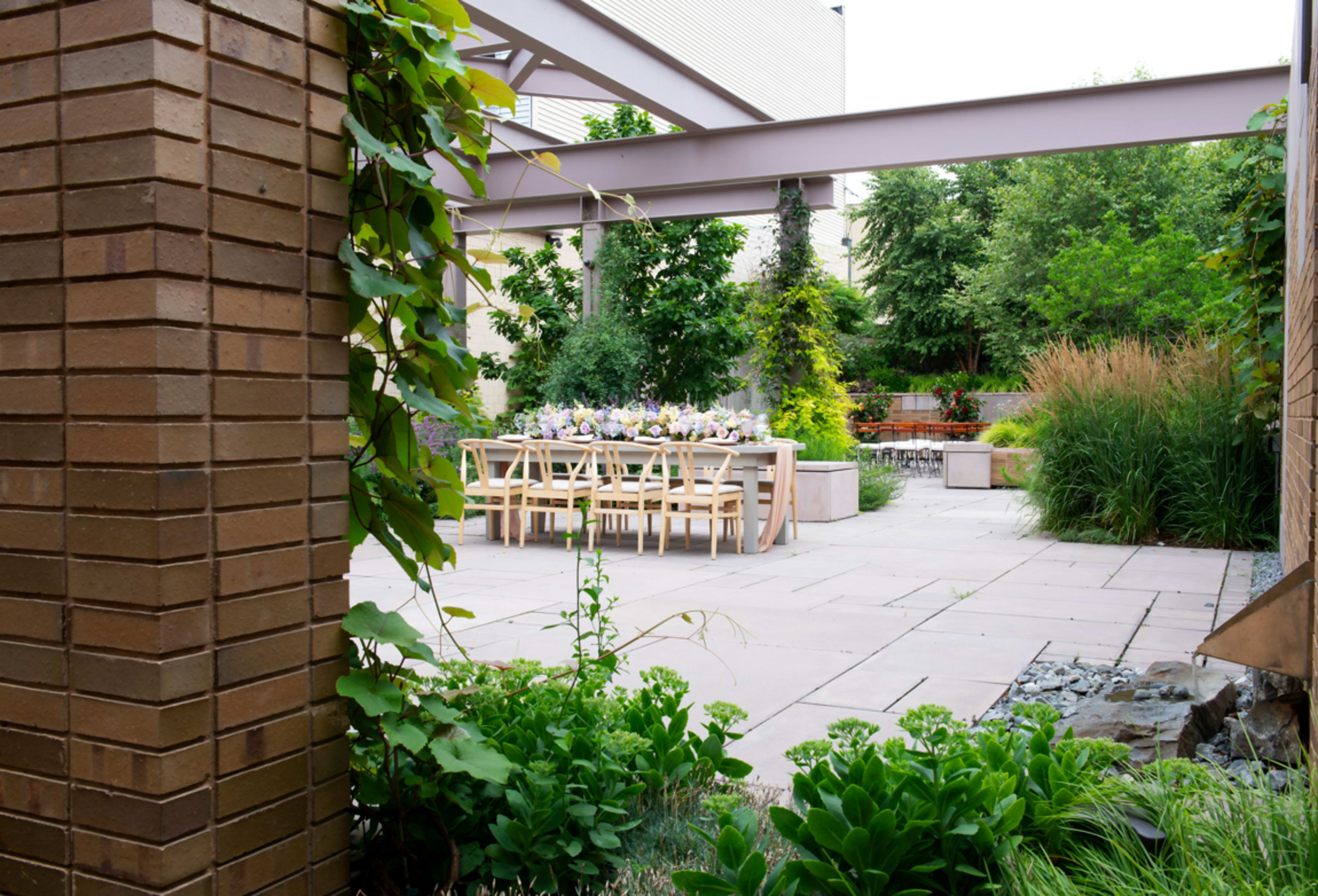 A stone patio with a long wooden dining table set for a meal is surrounded by greenery and garden features.