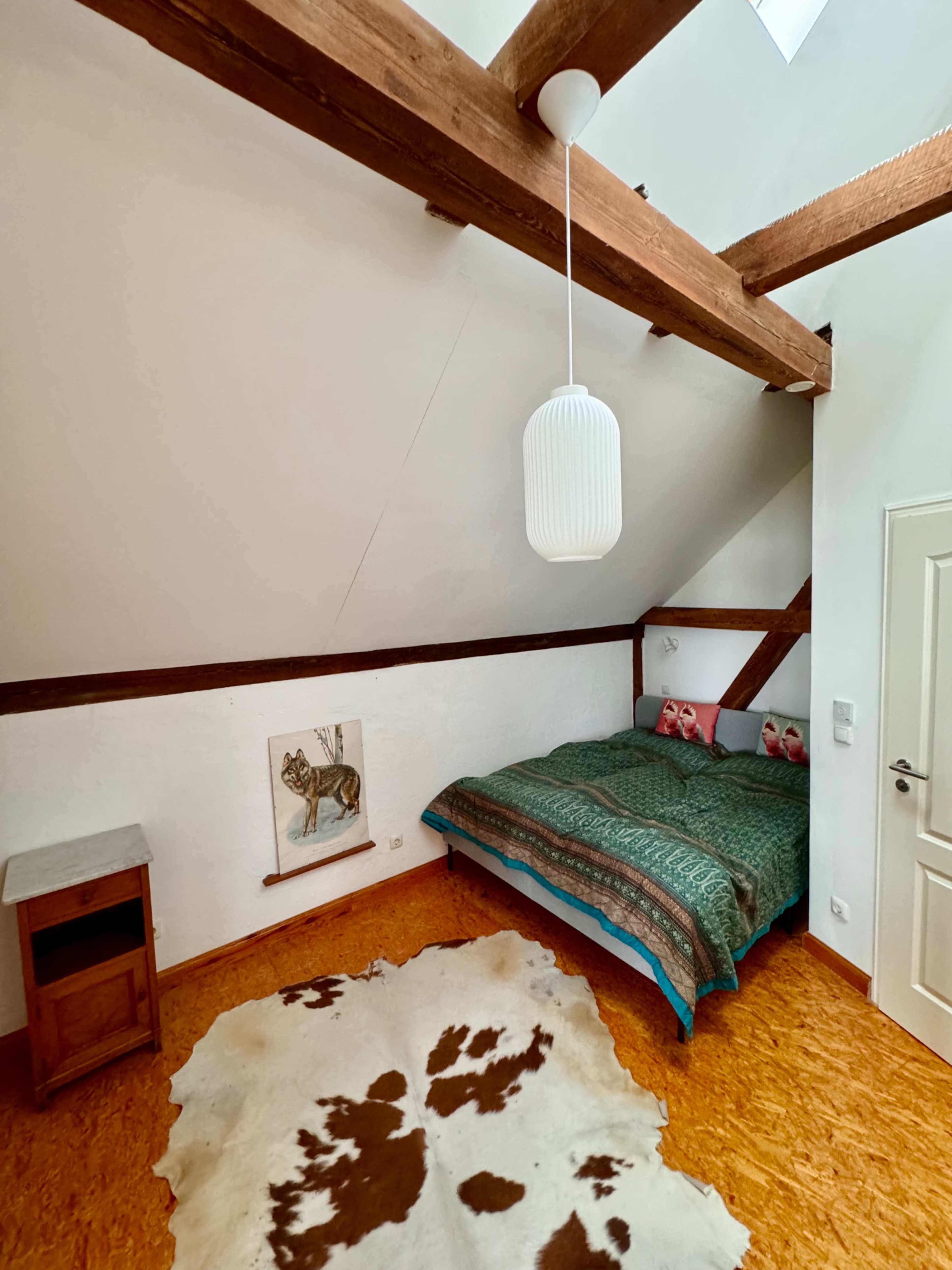 The image shows a cozy bedroom with a slanted ceiling, a wooden bed adorned with a patterned blanket, and a decorative cowhide rug on the floor.