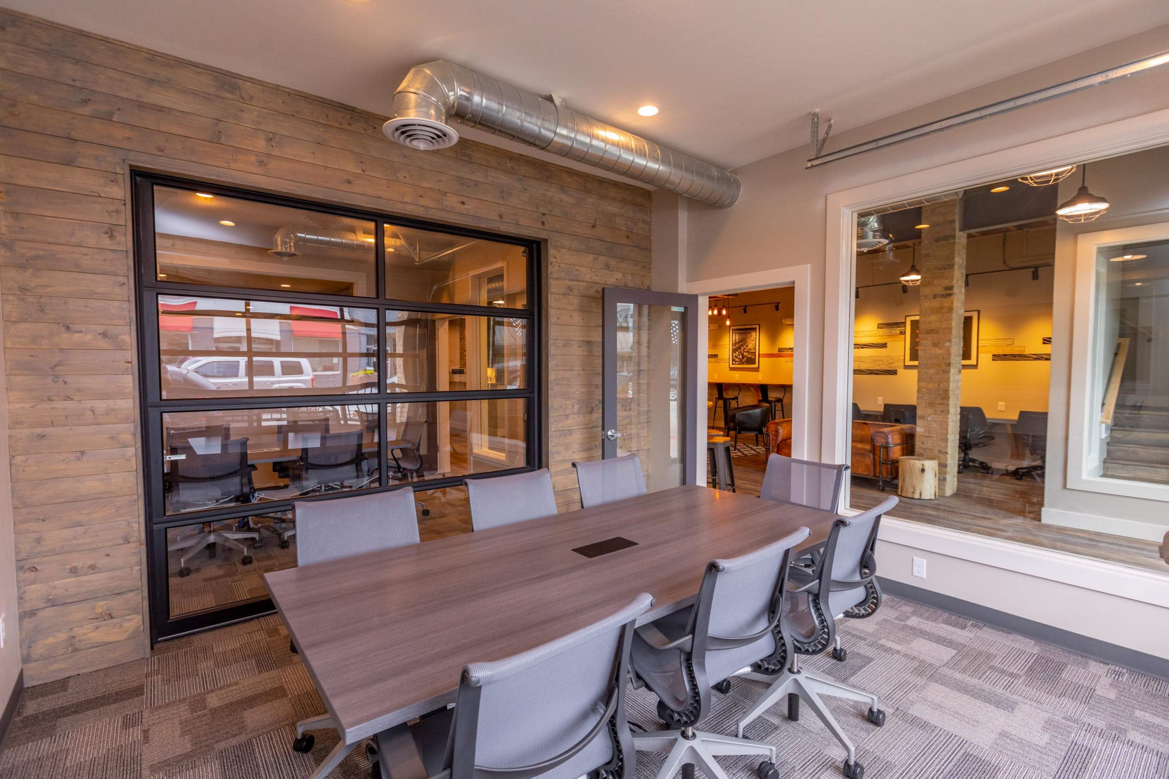 The image shows a modern conference room with a large table surrounded by chairs and a glass wall that provides a view into an adjoining meeting space.