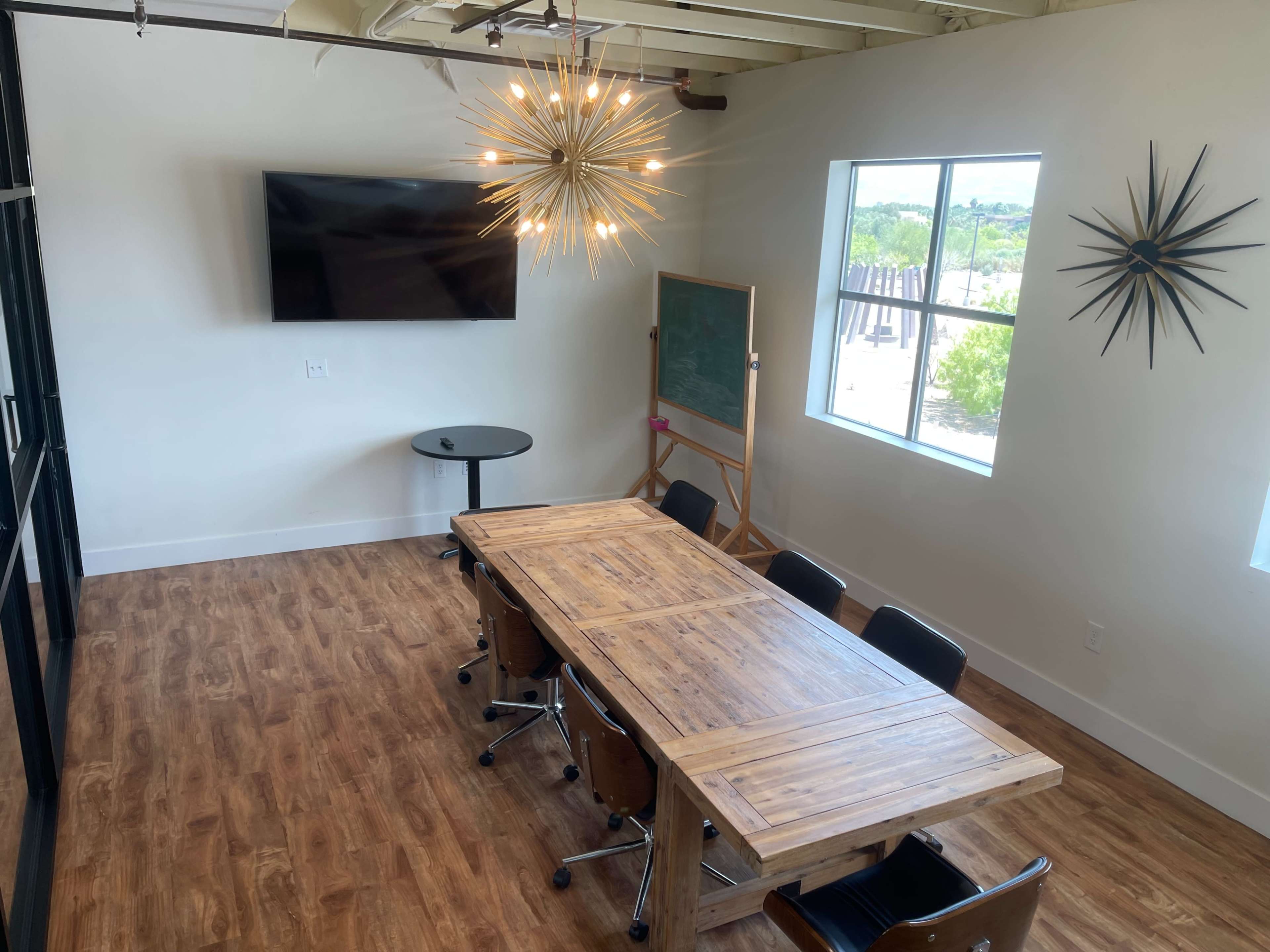 A modern conference room with a long wooden table, black chairs, a wall-mounted television, and a large window.