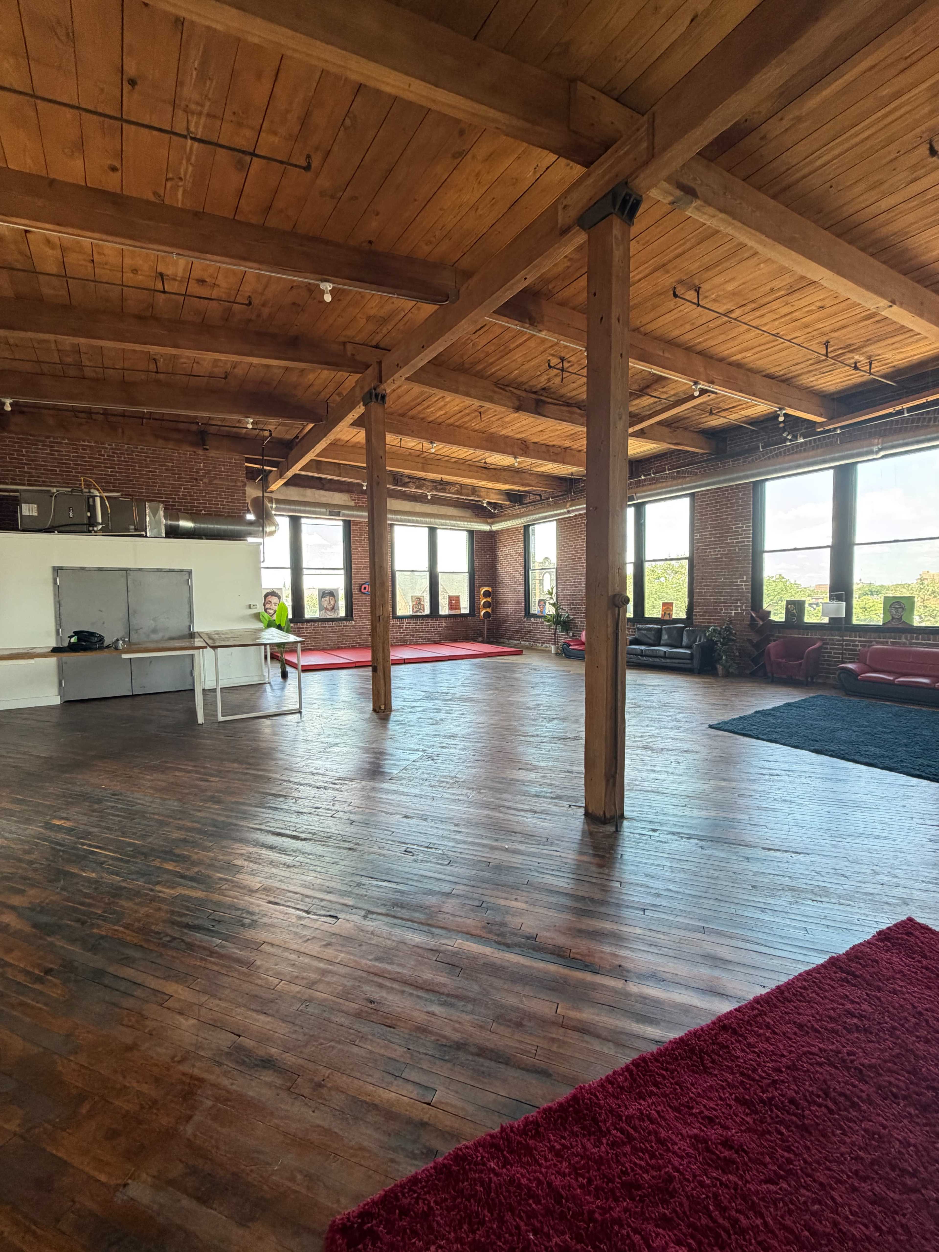 The image shows a spacious, open room with wooden floors, exposed beams, and large windows, featuring a kitchen area and a few seating arrangements.