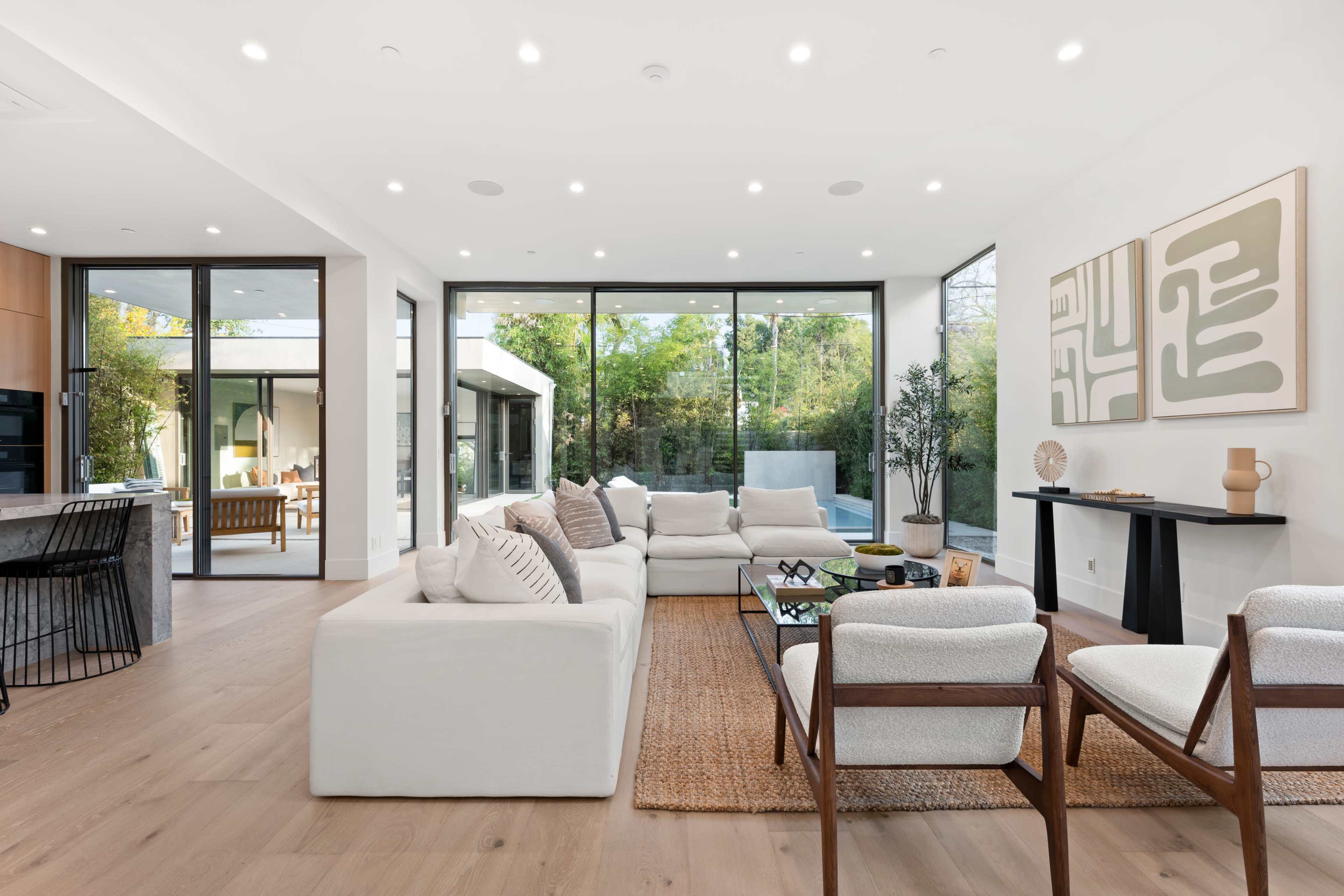 The modern living room features a large sectional sofa, minimalist furniture, and floor-to-ceiling windows that overlook a garden.