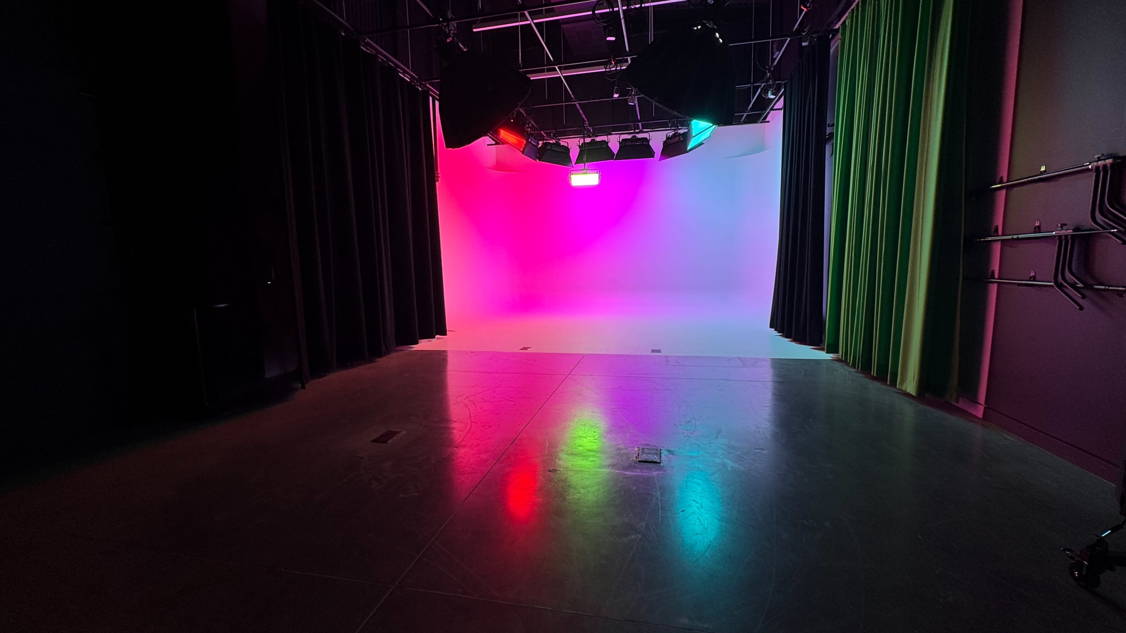 The image shows a studio space with a gradient backdrop of pink and blue lights, flanked by black and green curtains.