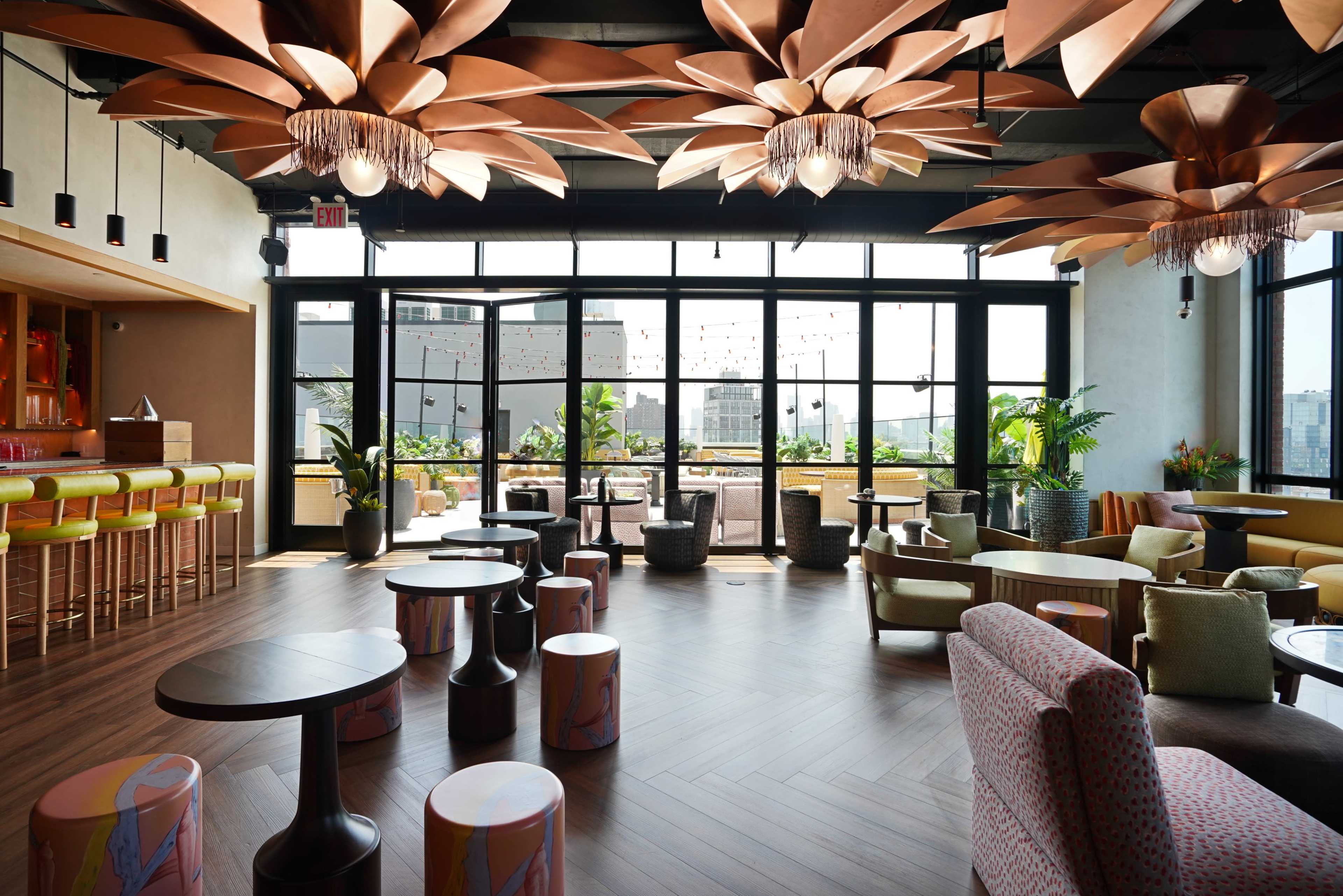 The image shows a modern rooftop lounge with large windows, stylish furniture, and decorative pendant lights resembling flowers.