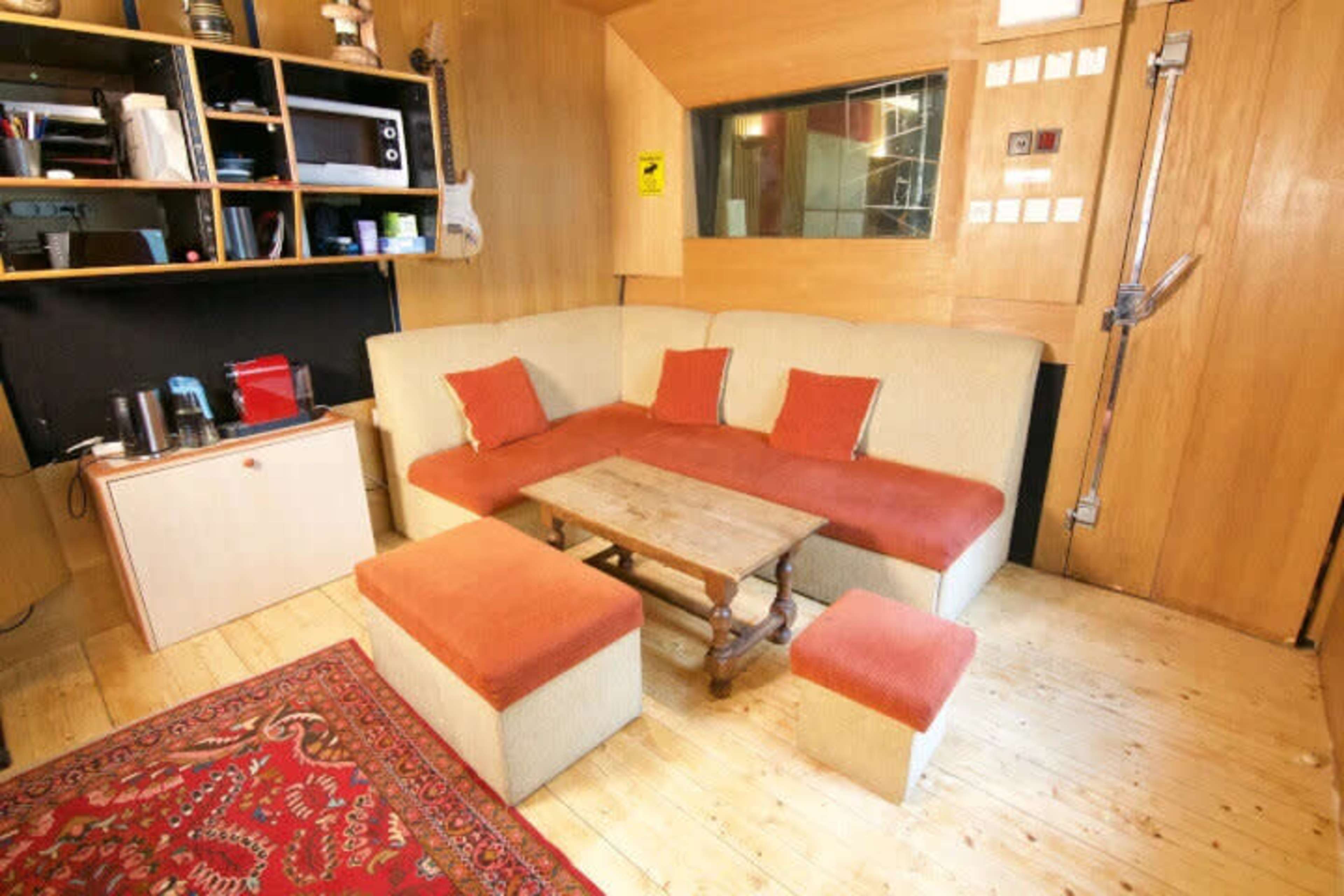The image shows a cozy wooden room with a sofa, a coffee table, and a small area for appliances along with shelves and a carpet.