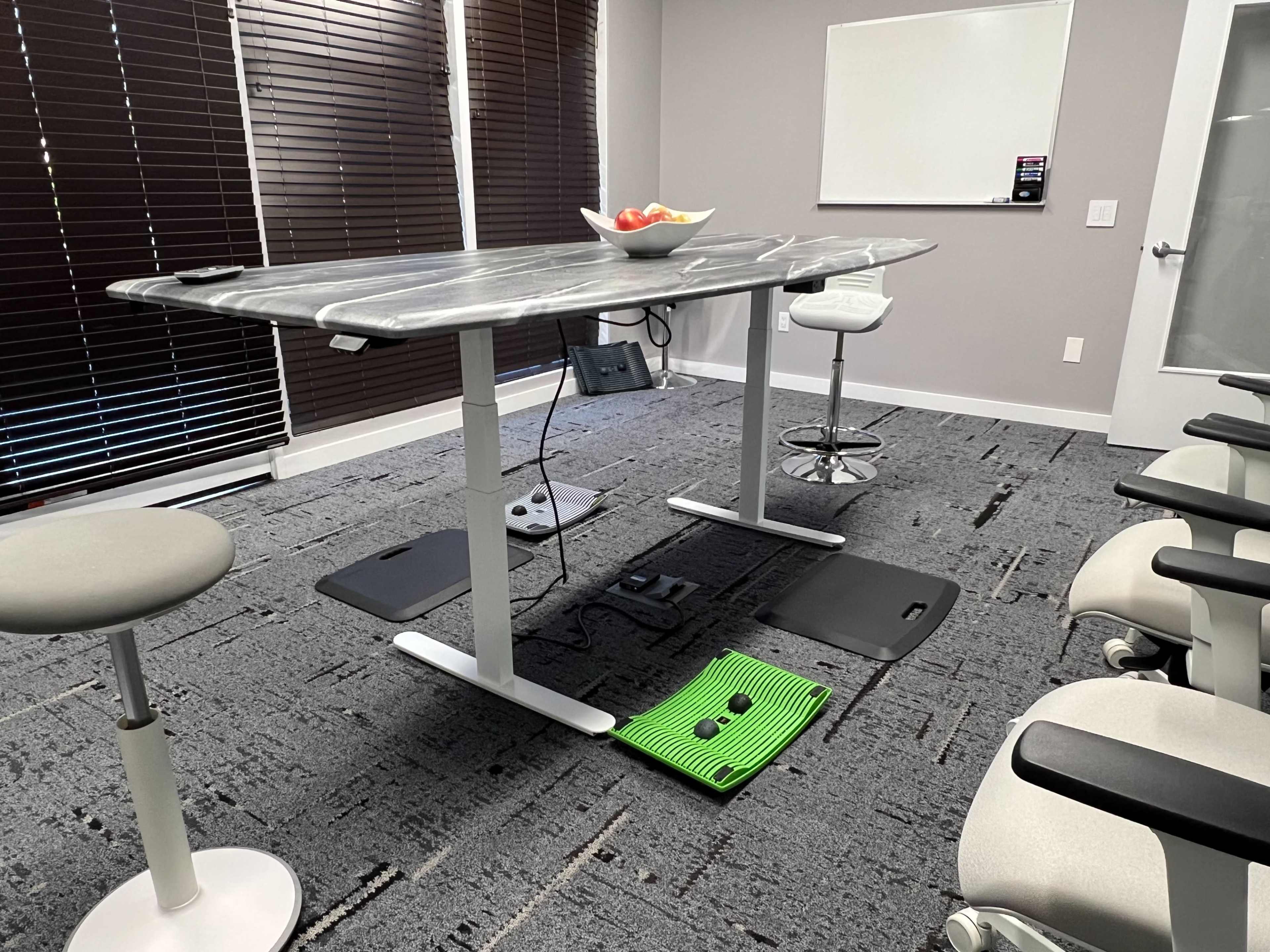 A modern meeting room features a height-adjustable table with a bowl of fruit on top, surrounded by ergonomic chairs and a green mat on the floor.