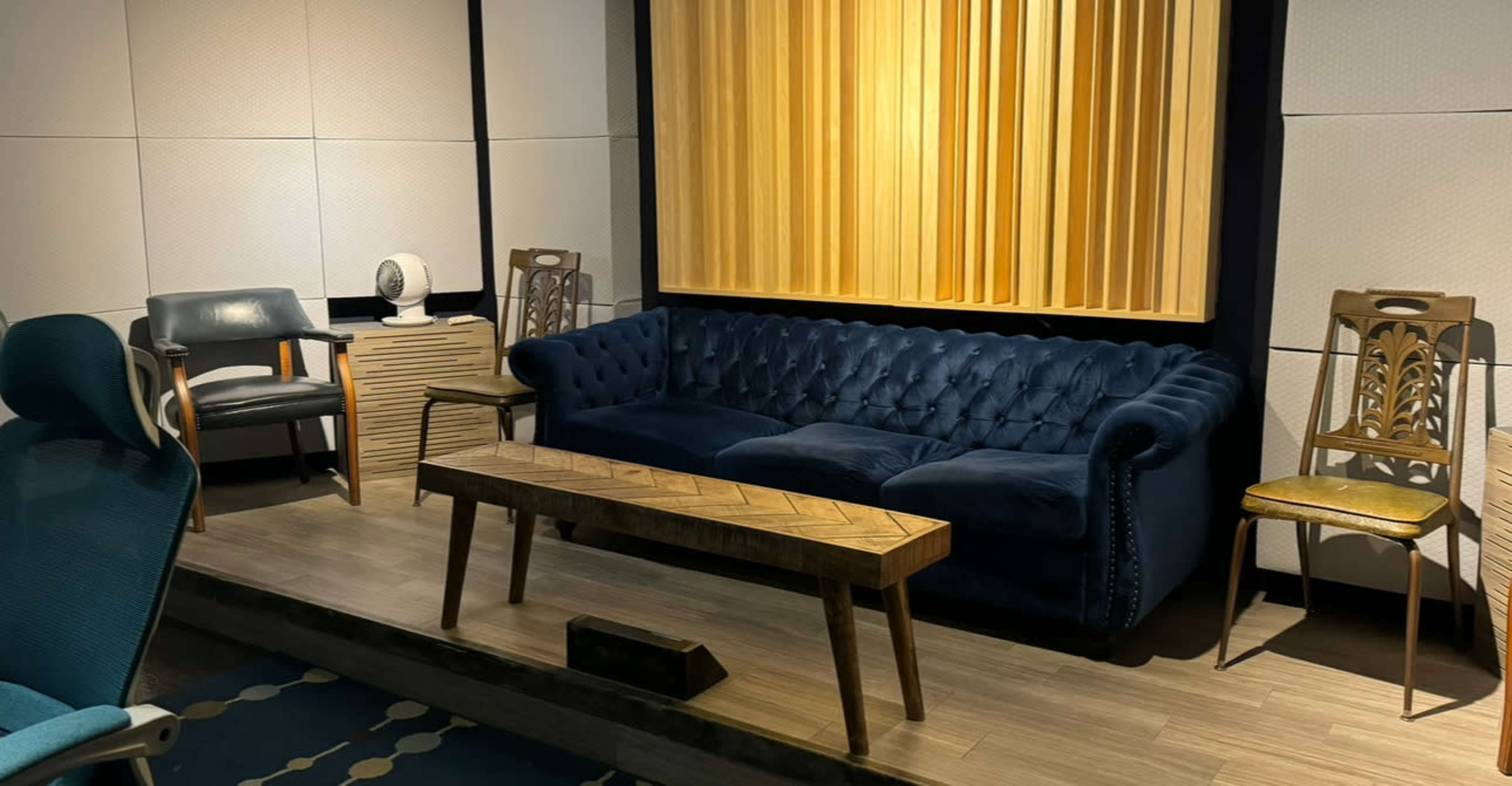 The image shows a modern interior with a blue velvet sofa, a wooden coffee table, and two chairs, set against a backdrop of acoustic panels.