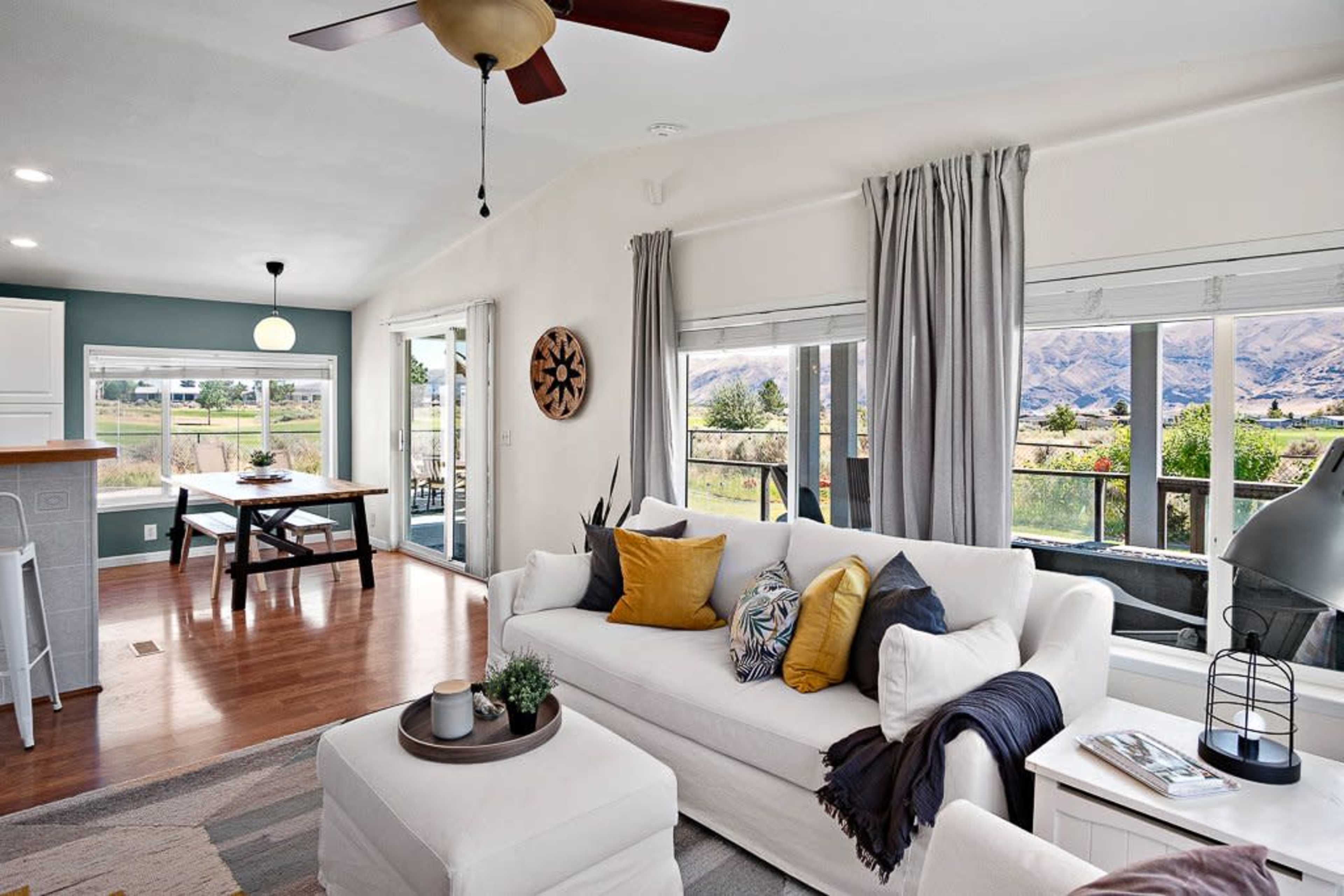 A bright living room features a white sofa adorned with decorative pillows, large windows with views of a natural landscape, and a dining area in the background.