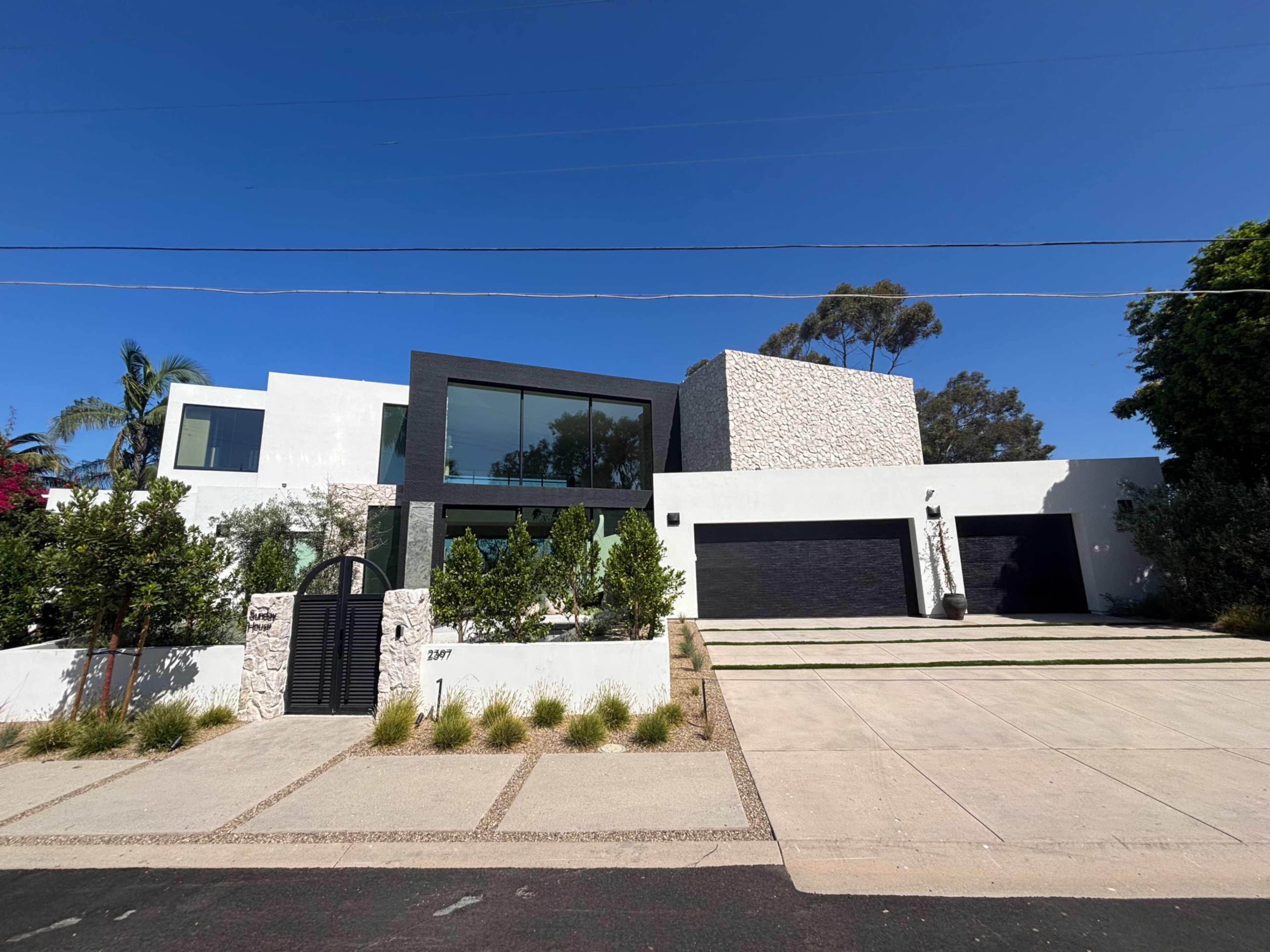 The image shows a modern, two-story house with a white exterior and black accents, featuring a landscaped front yard and a driveway with a double garage.