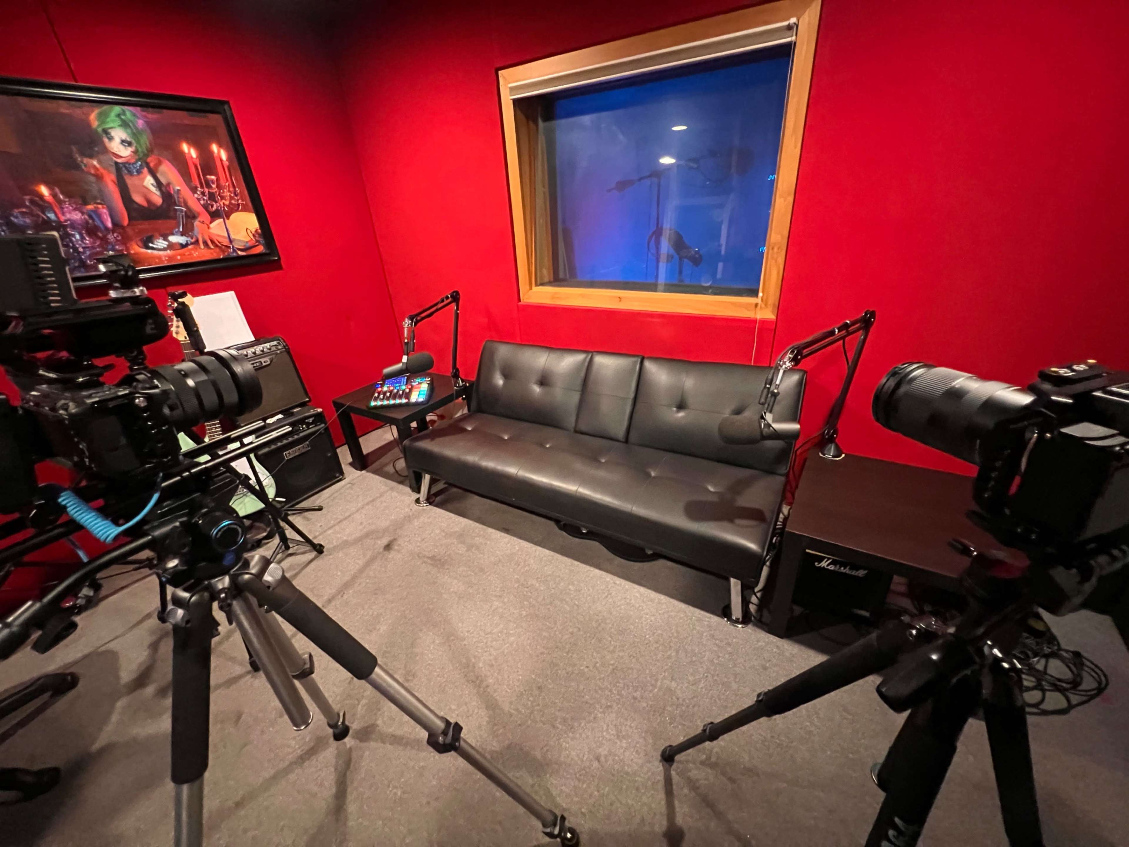 The image shows a recording studio with a red wall, two cameras mounted on tripods facing a black sofa, and a window revealing another room with blue lighting.