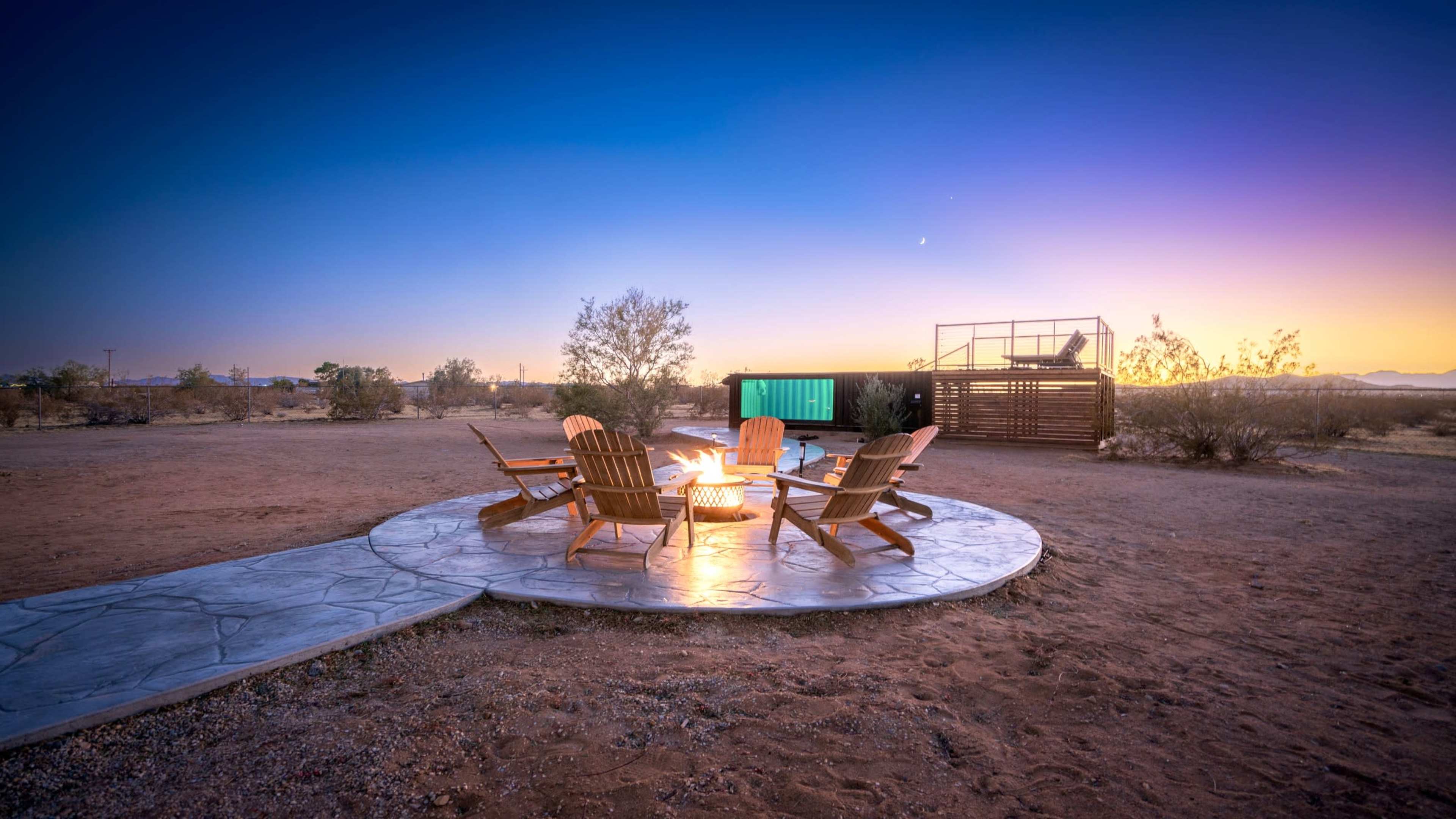A circular stone patio with wooden chairs surrounds a fire pit, set against a desert landscape at dusk with vibrant colors in the sky.
