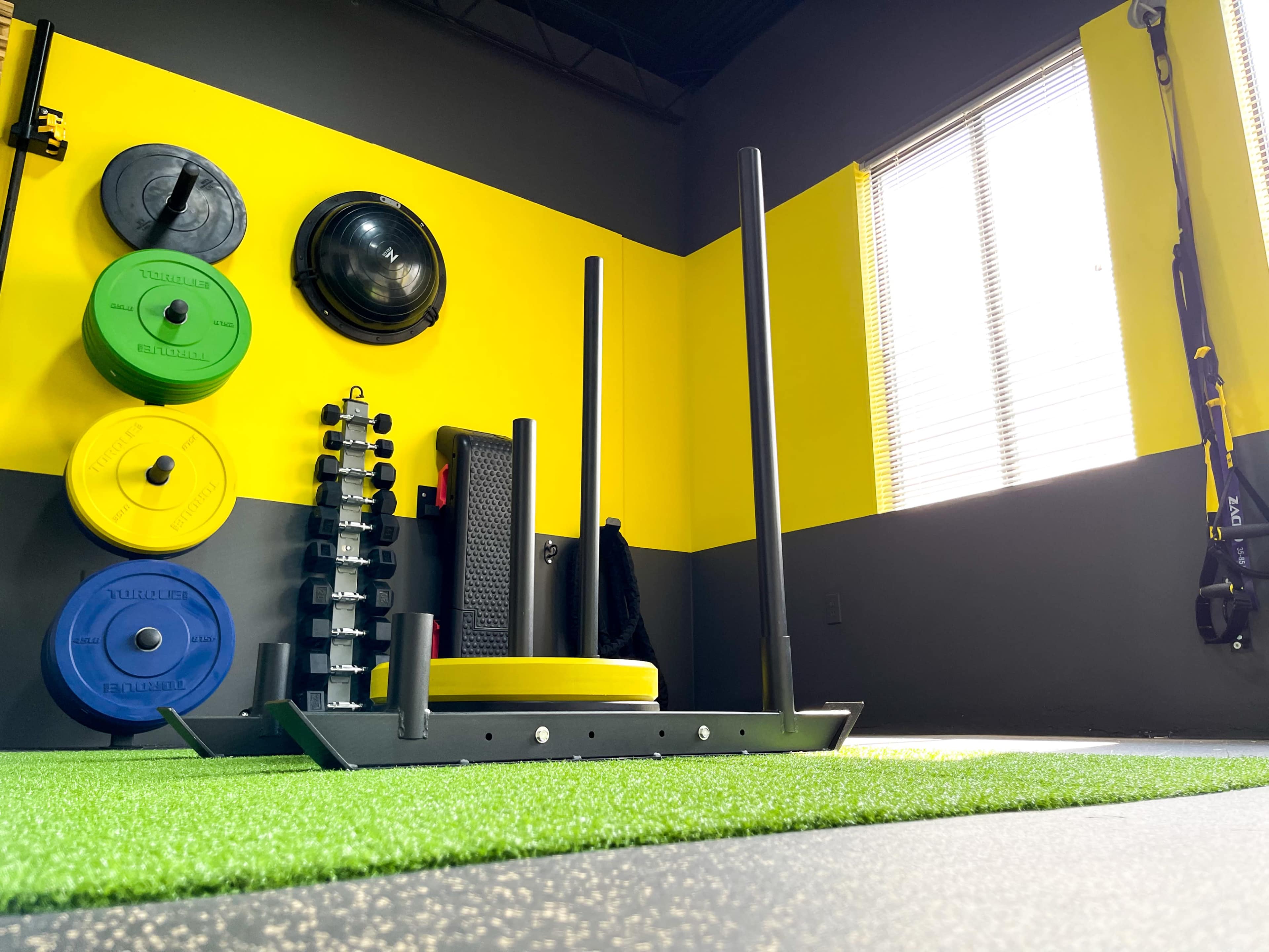 A functional gym space with a yellow and black color scheme, featuring a sled, weight plates, and exercise equipment against a green turf floor.