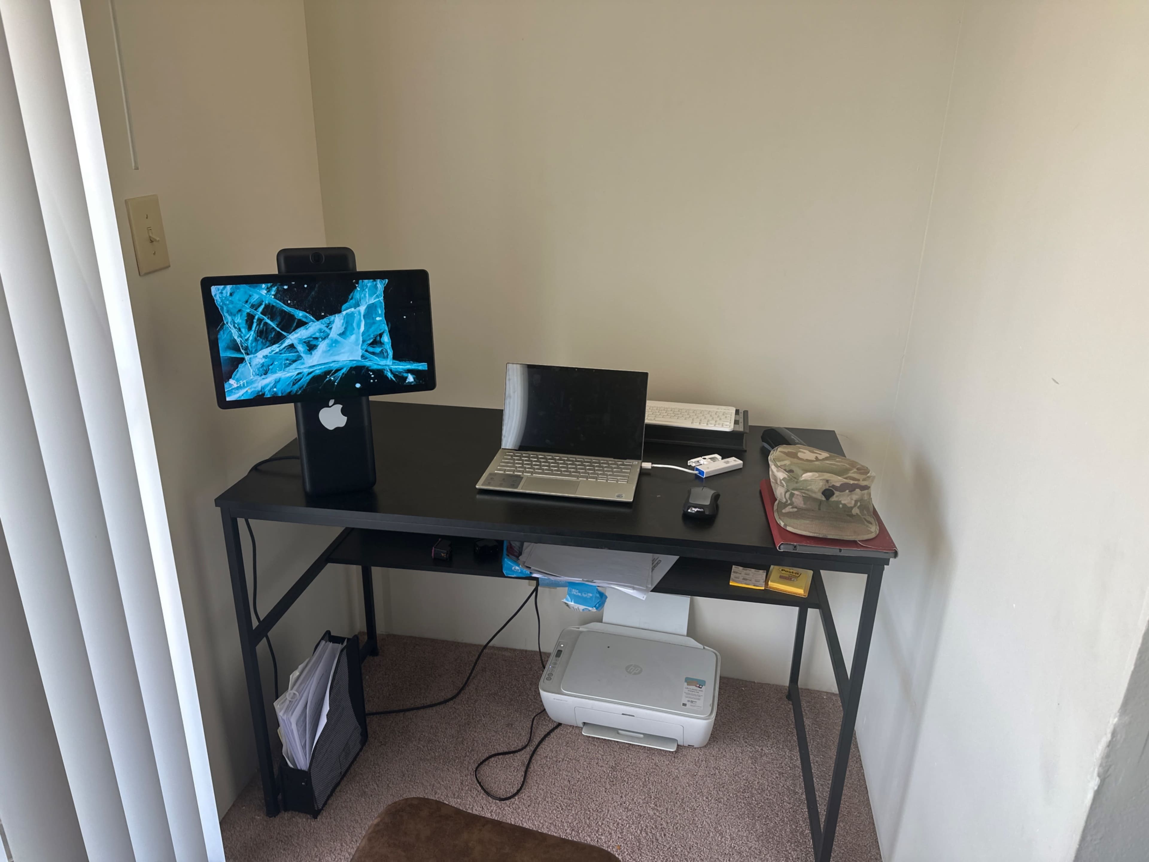 The image shows a compact desk setup in a corner with a desktop monitor, a laptop, a printer, and a few office supplies.