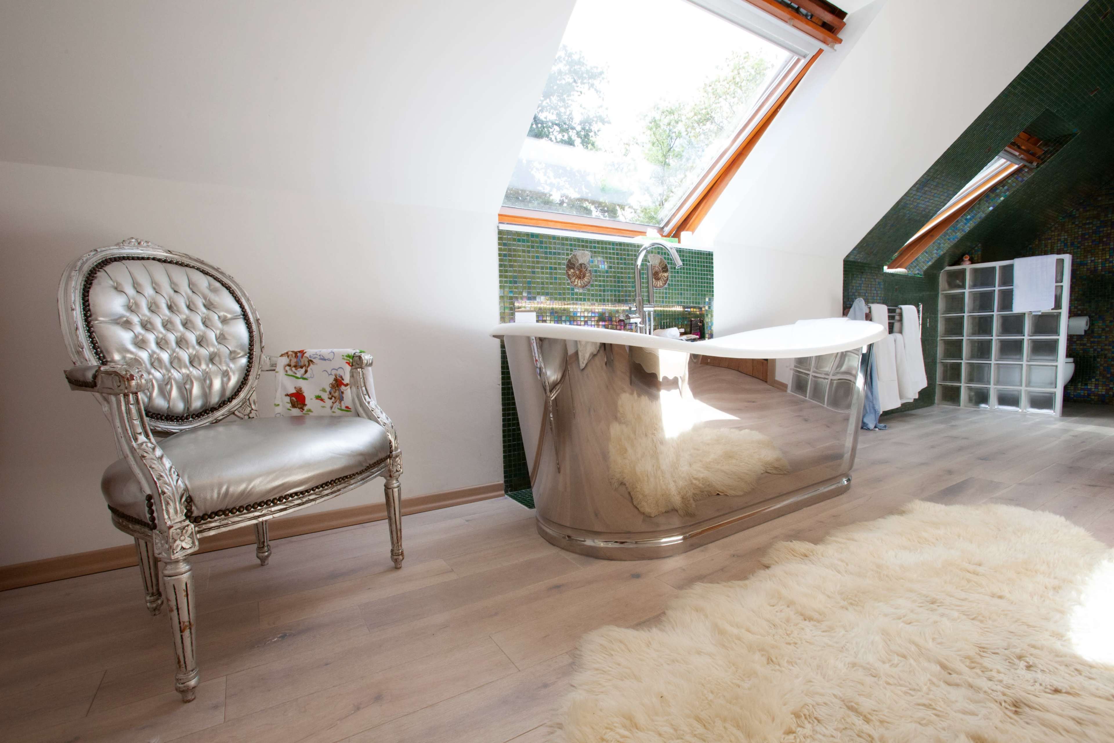 A modern bathroom with a sleek, reflective bathtub, a silver chair, and natural light streaming in through skylights.