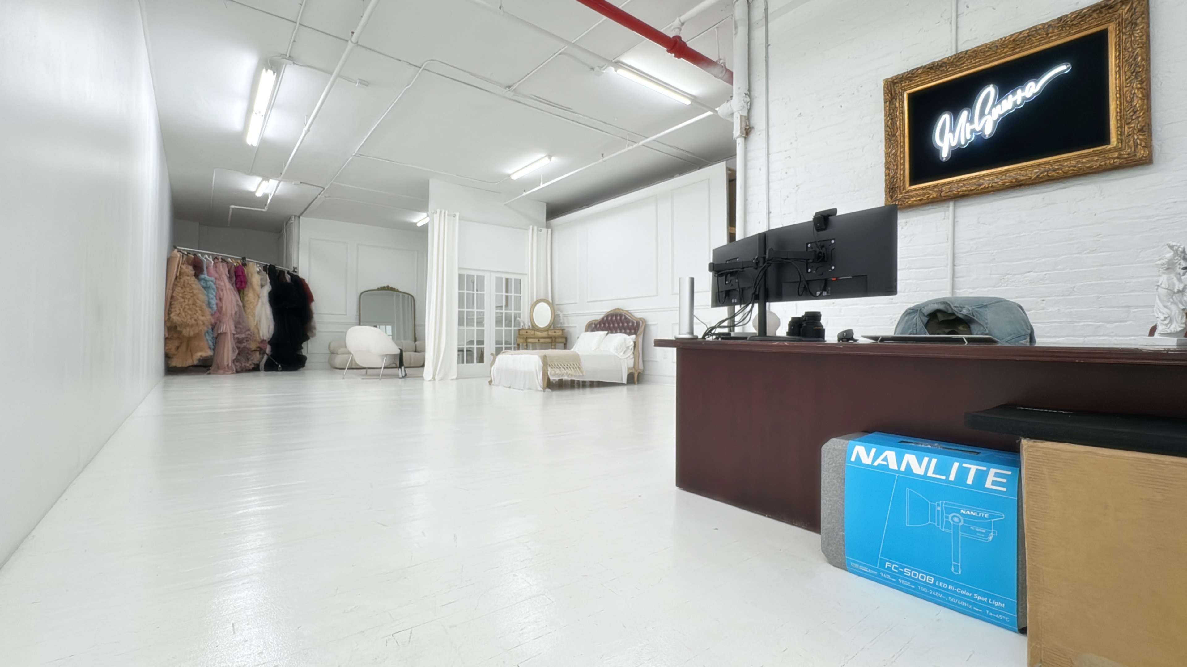 The image shows a bright, spacious studio or office area with white walls and flooring, featuring a desk with a computer setup, several pieces of furniture, and a clothing rack in the background.