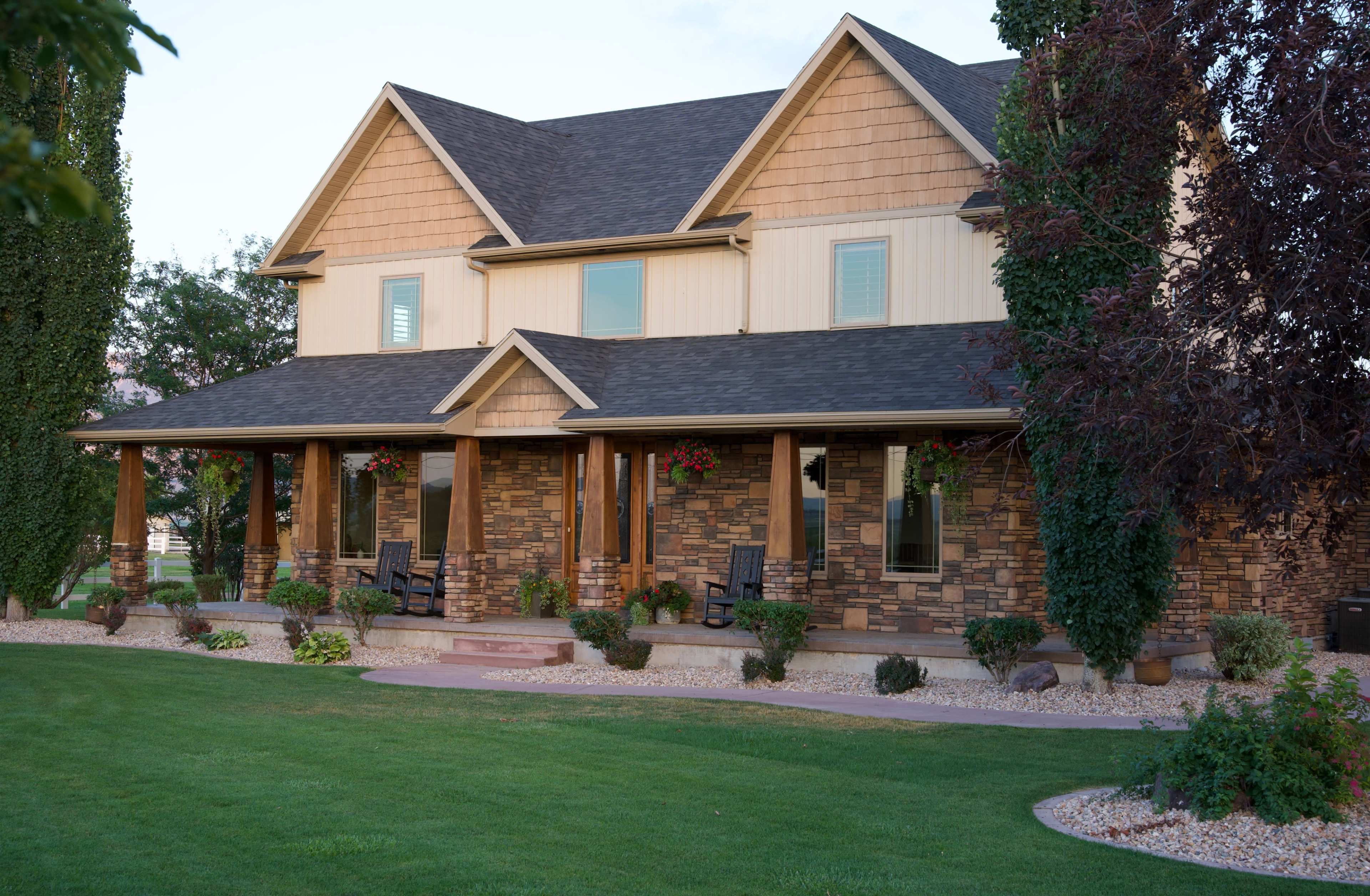 A two-story house features a stone facade, gabled roof, and a landscaped front yard with flower beds and seating areas.