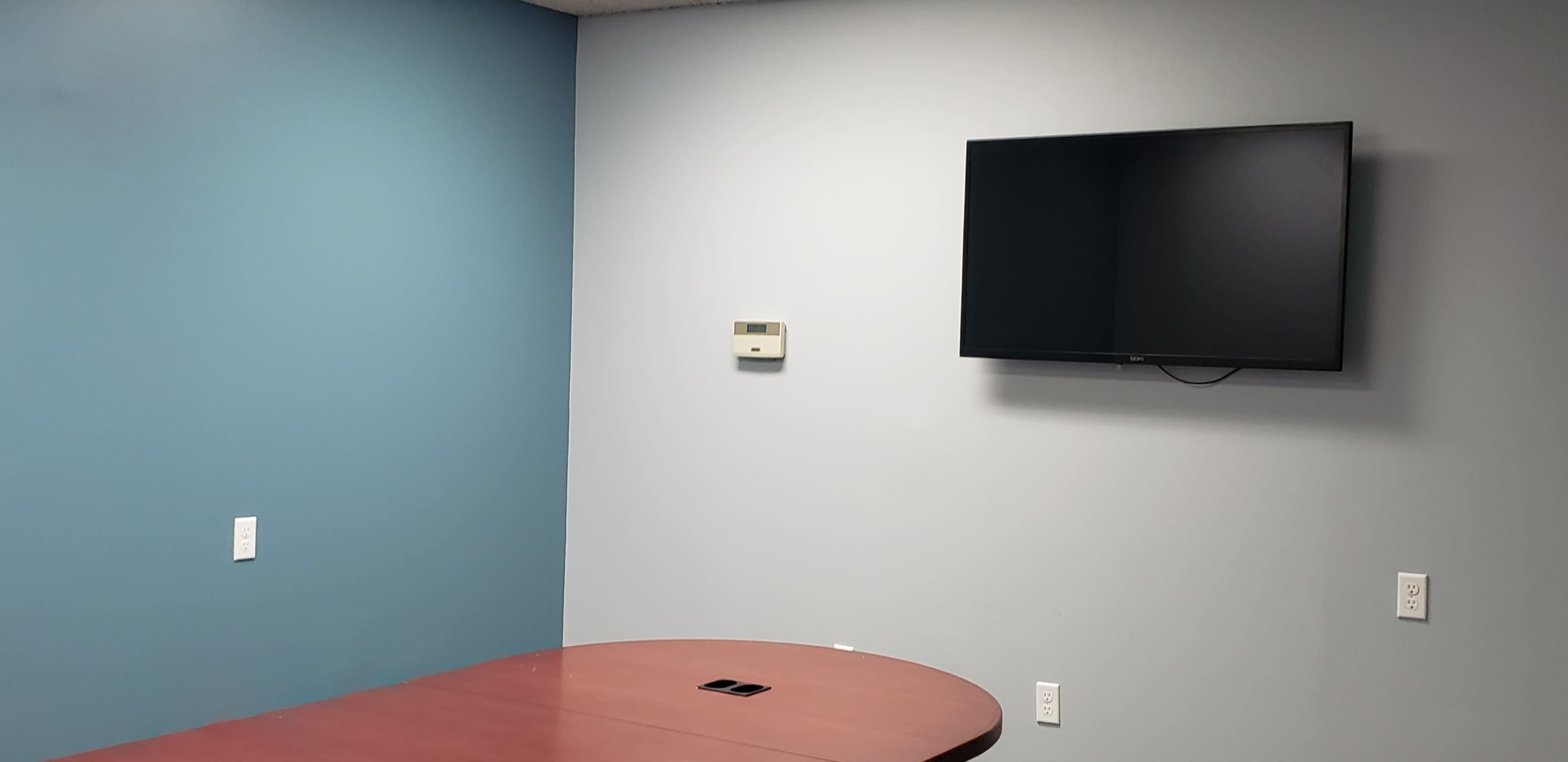 A conference room with a brown oval table, a wall-mounted TV, and blue and gray walls.