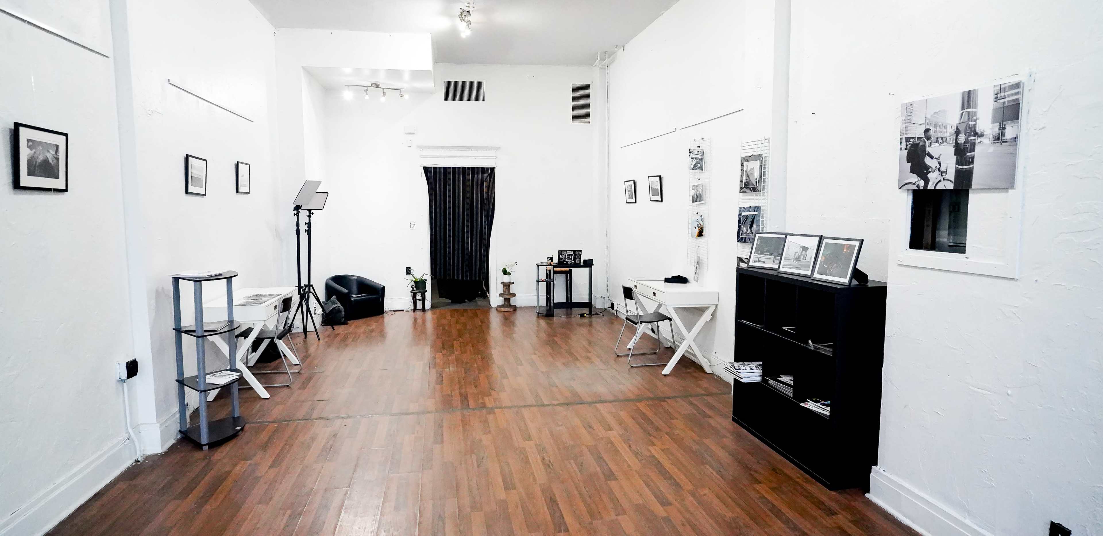 A spacious room with wooden flooring, white walls, and several framed photographs displayed on the walls, along with furniture including tables and a chair.
