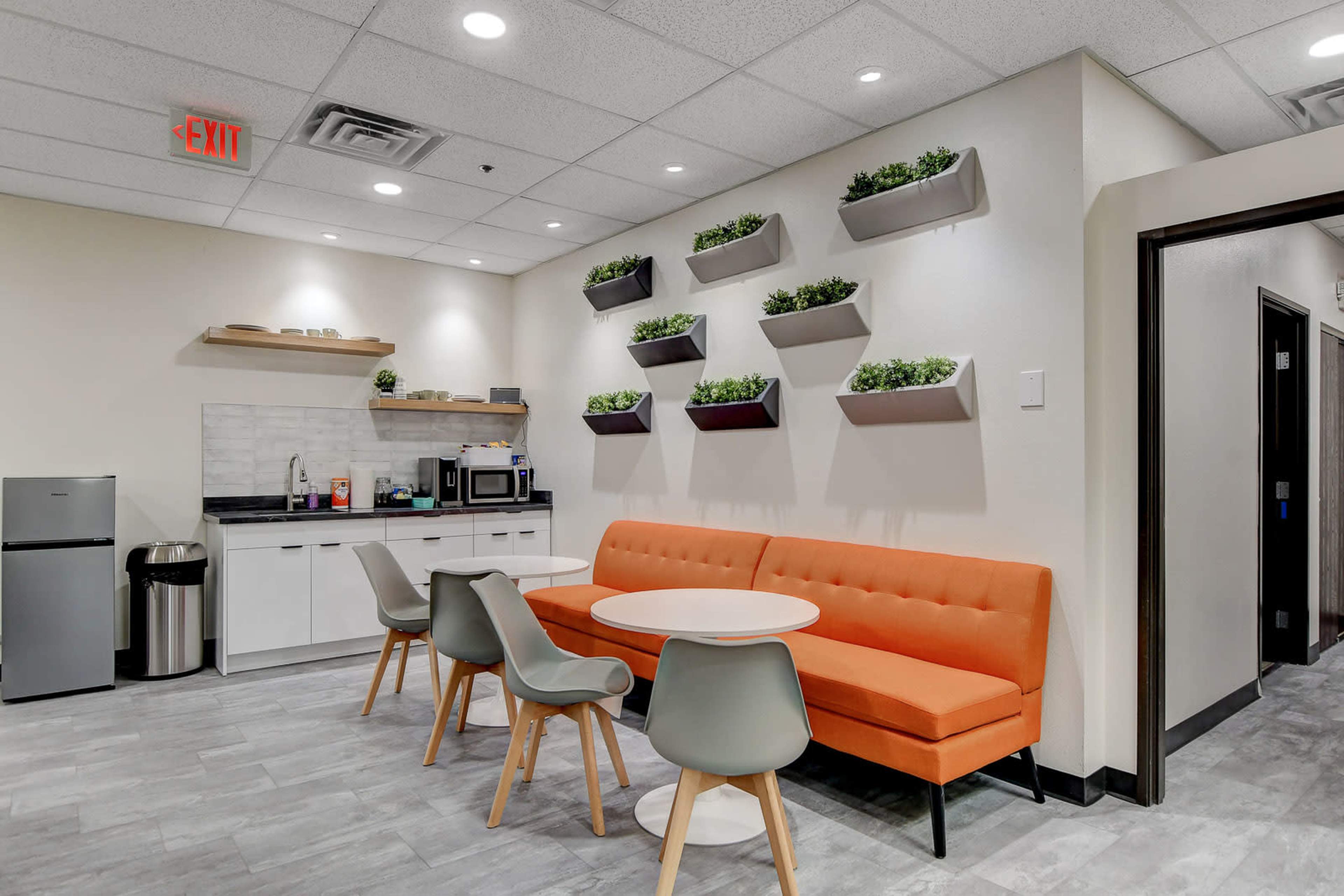 The image shows a modern break room with an orange sofa, a circular table, and wall-mounted planters, along with kitchen appliances and a refrigerator.
