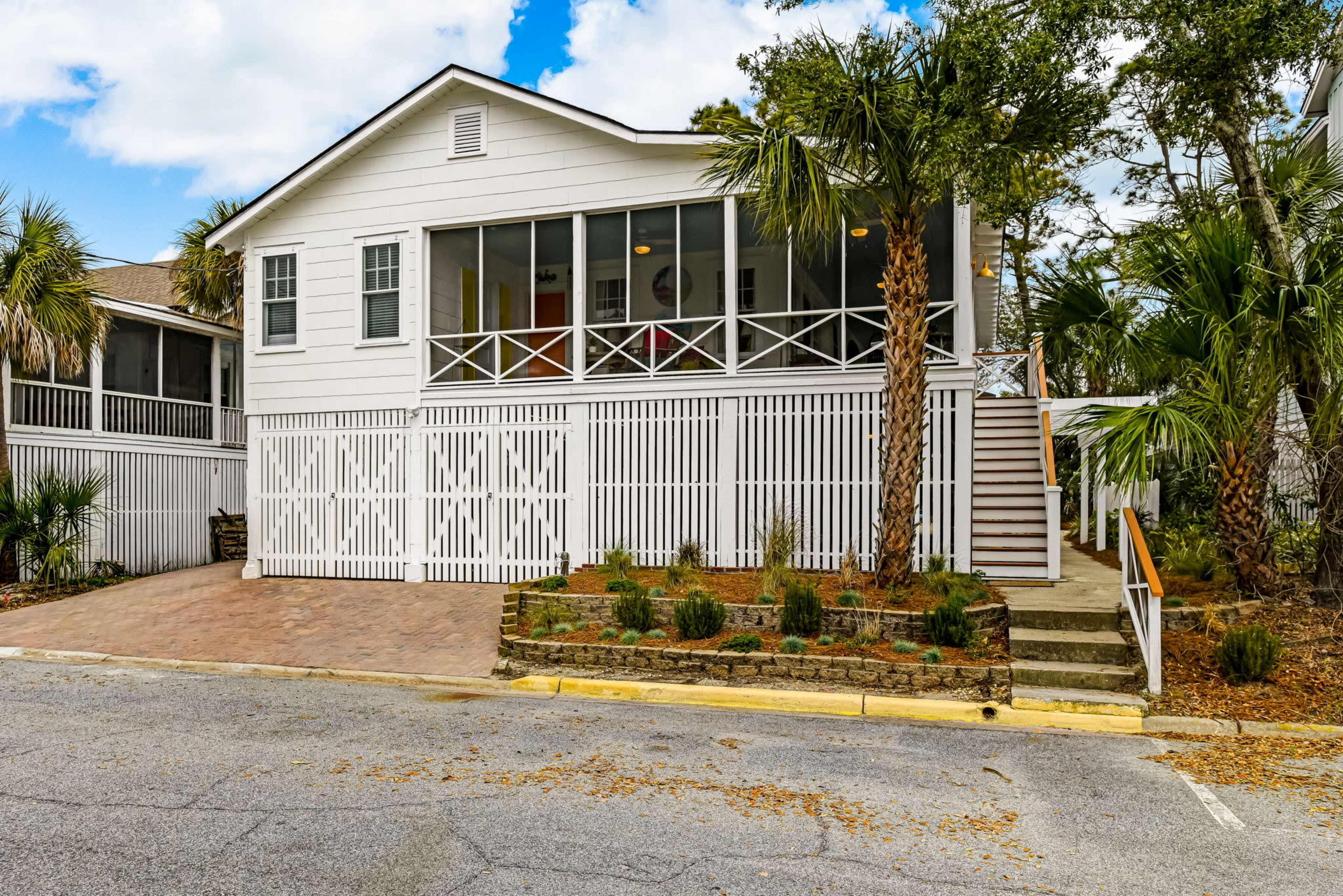 A two-story white house with a screened porch and palm trees is located on a paved lot beside a street.