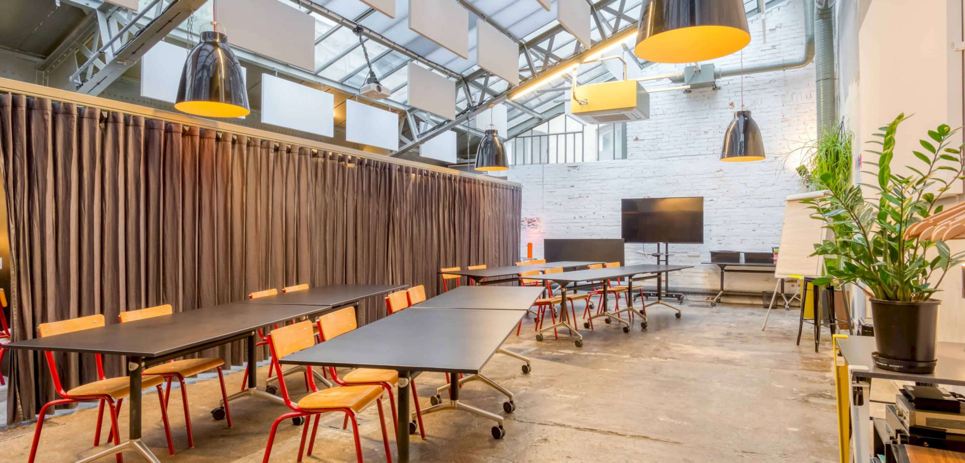 The image shows a modern conference room with multiple black tables, wooden chairs, and large industrial light fixtures, featuring gray curtains and a presentation screen at the back.