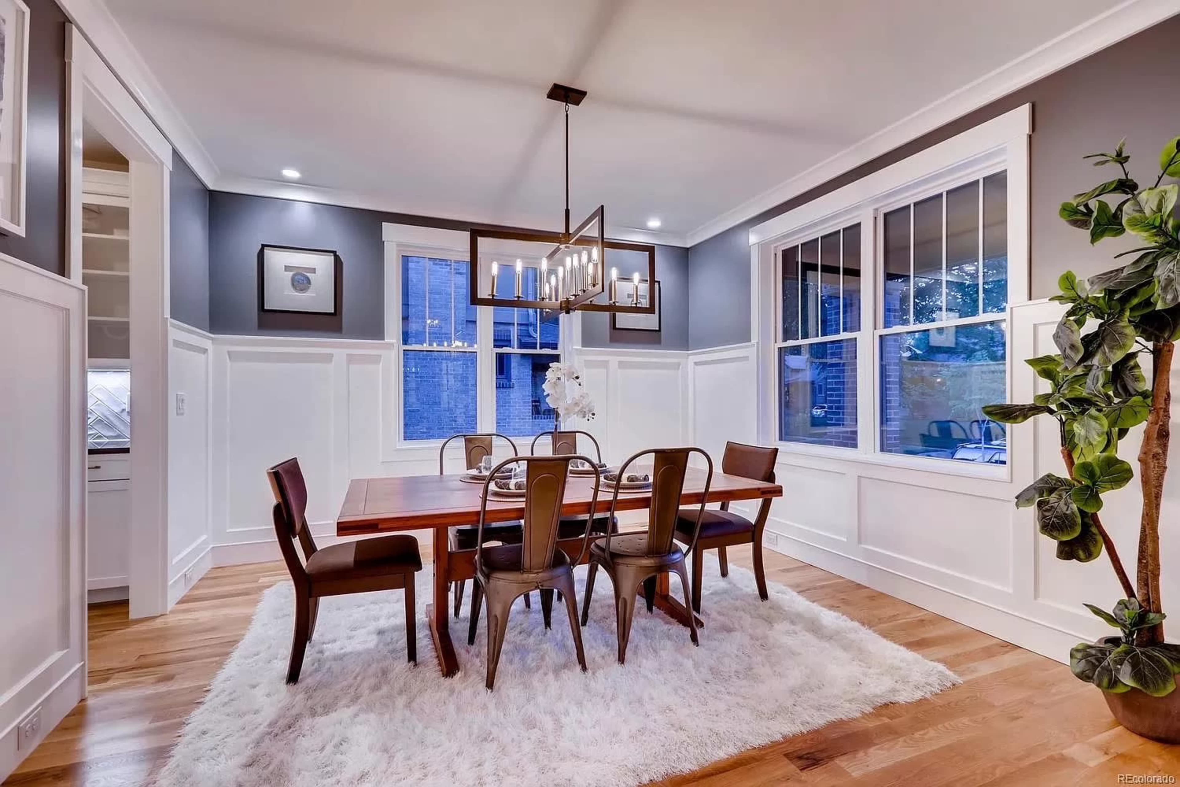 A dining room features a wooden table surrounded by metal chairs, with large windows and decorative wall paneling.