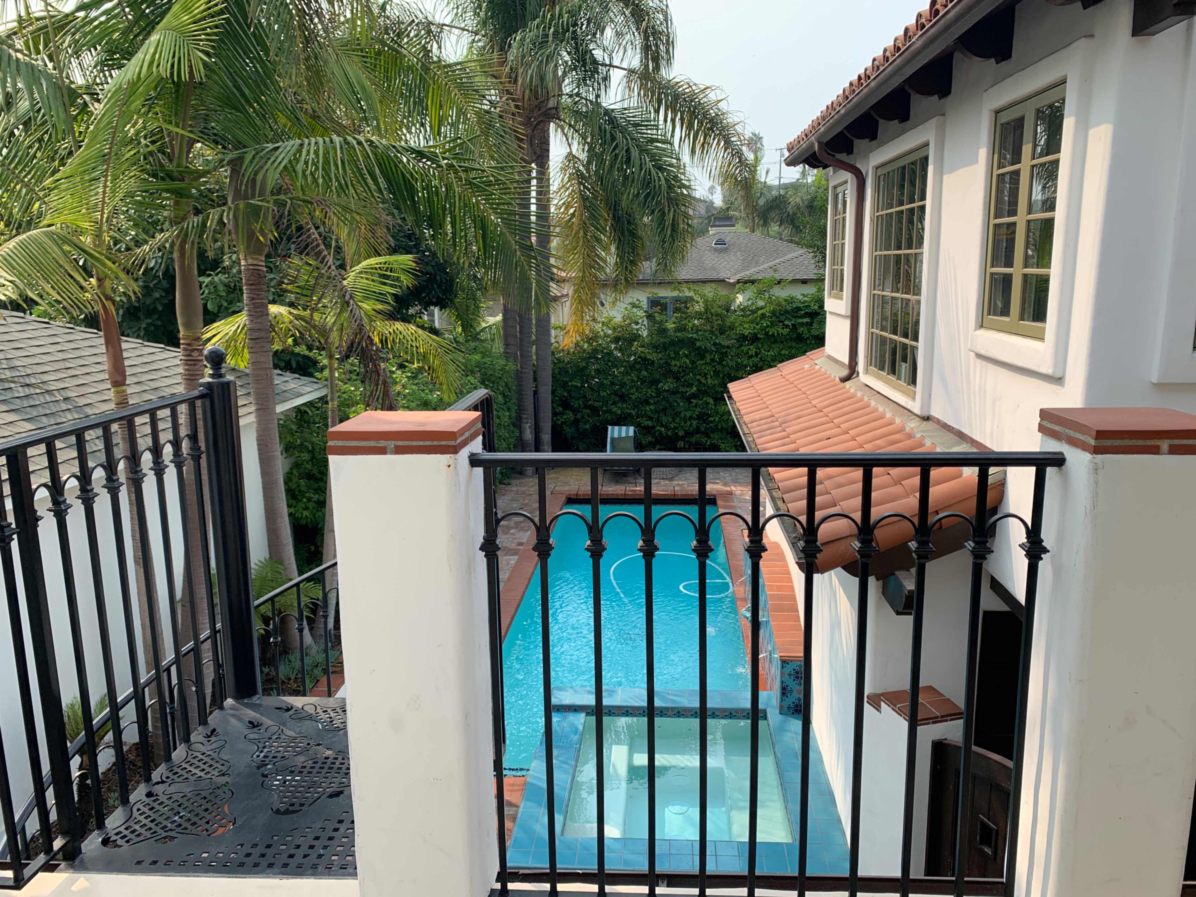 A view of a swimming pool surrounded by palm trees, seen from a balcony with a wrought iron railing.