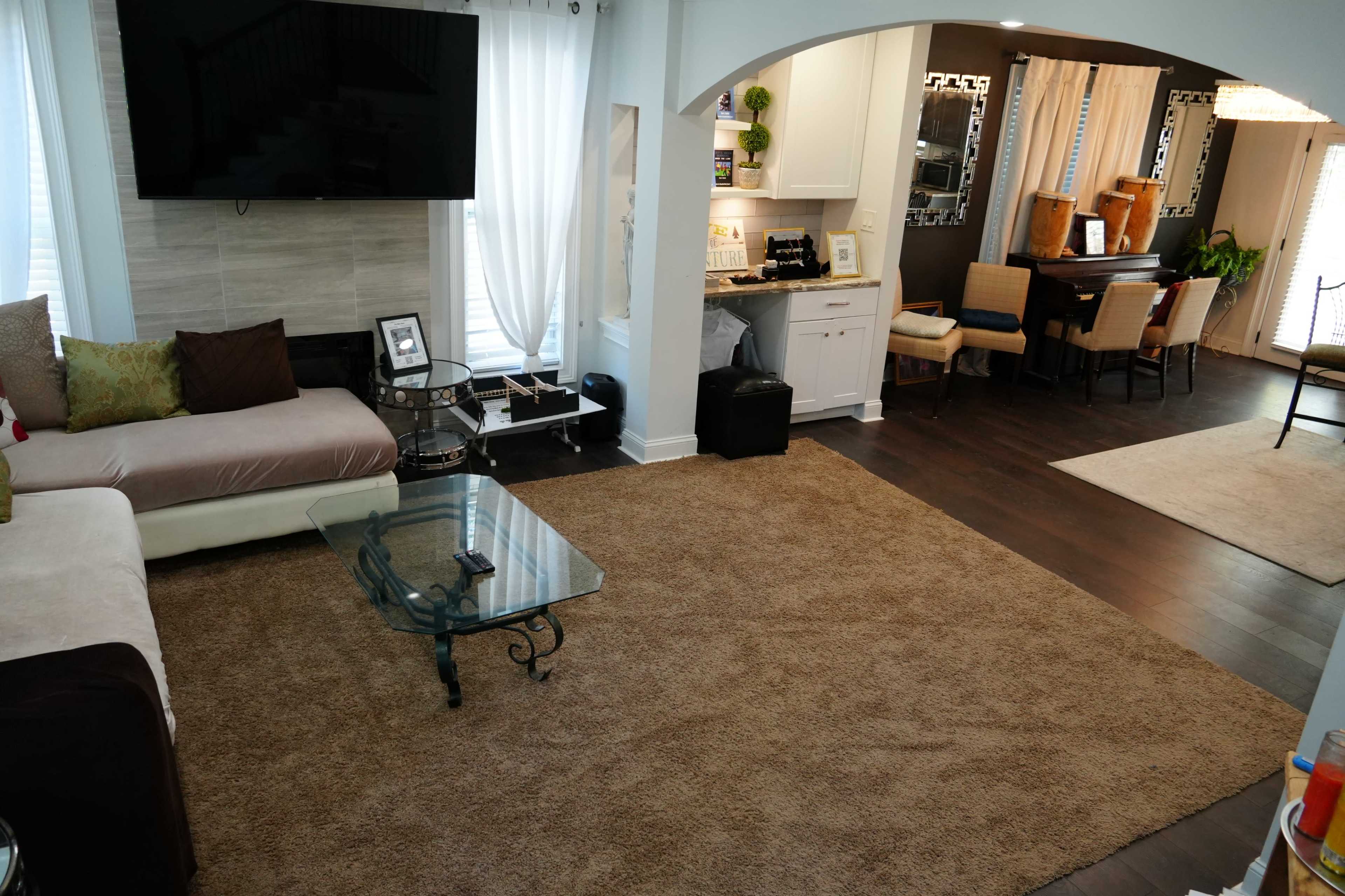 A living room with a large beige rug, a coffee table, a sectional sofa, and an open space connecting to a dining area with a table and chairs.